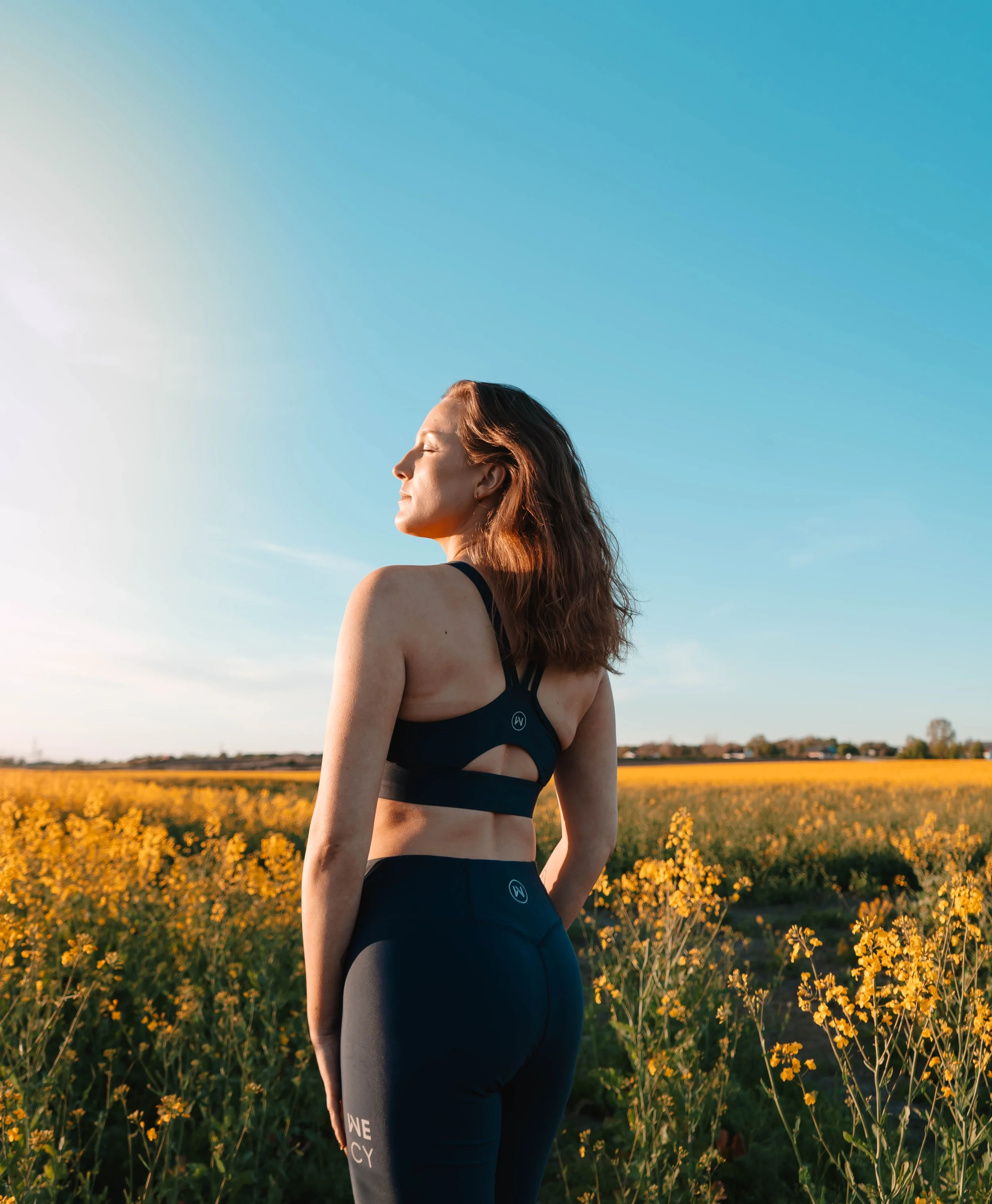 A woman with long brown hair stands with her eyes closed and face turned slightly upward in a field of yellow flowers under a clear blue sky during sunset.