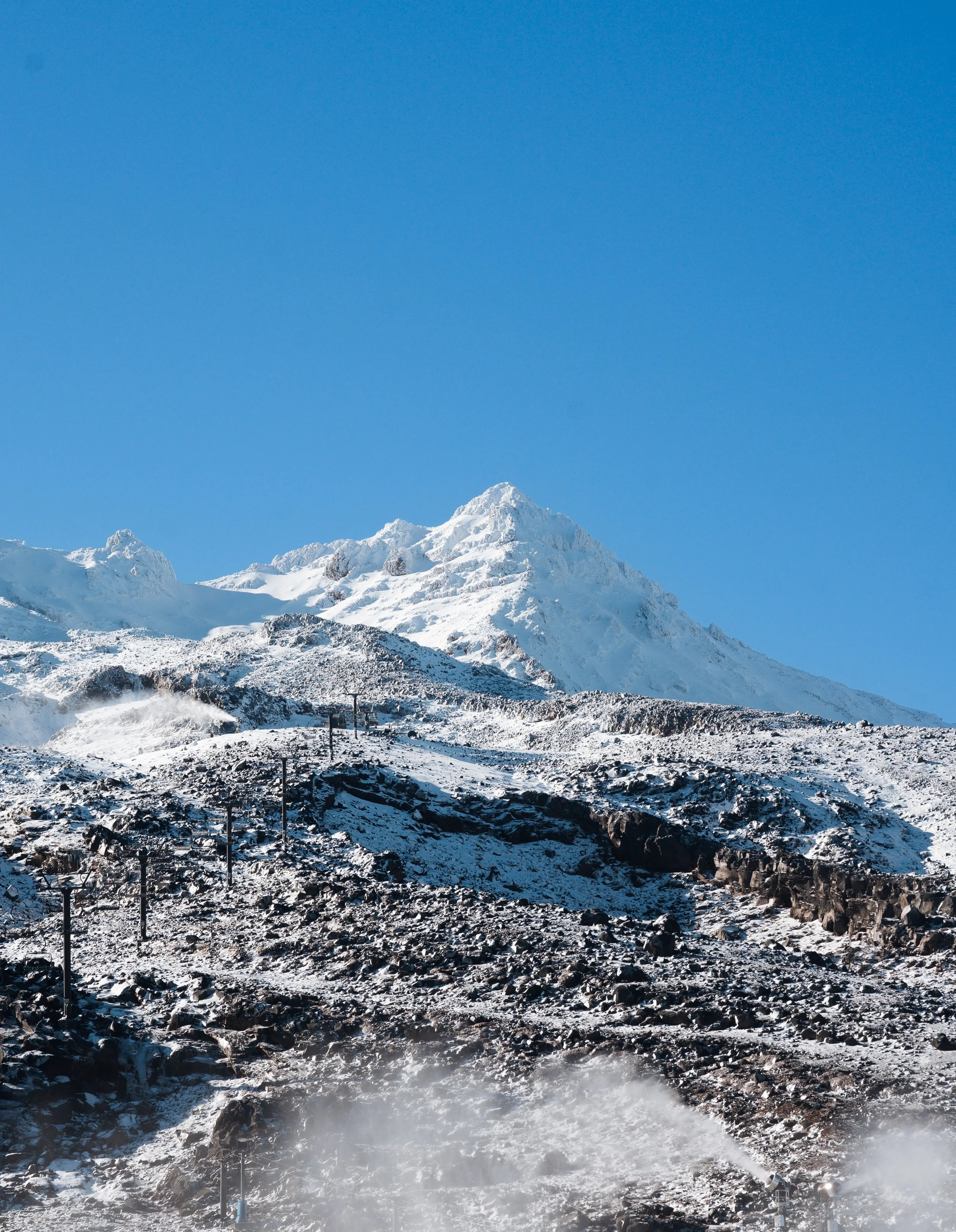 Snow-covered mountain with a clear blue sky, ski lifts, and steam or smoke rising from the rocky terrain.
