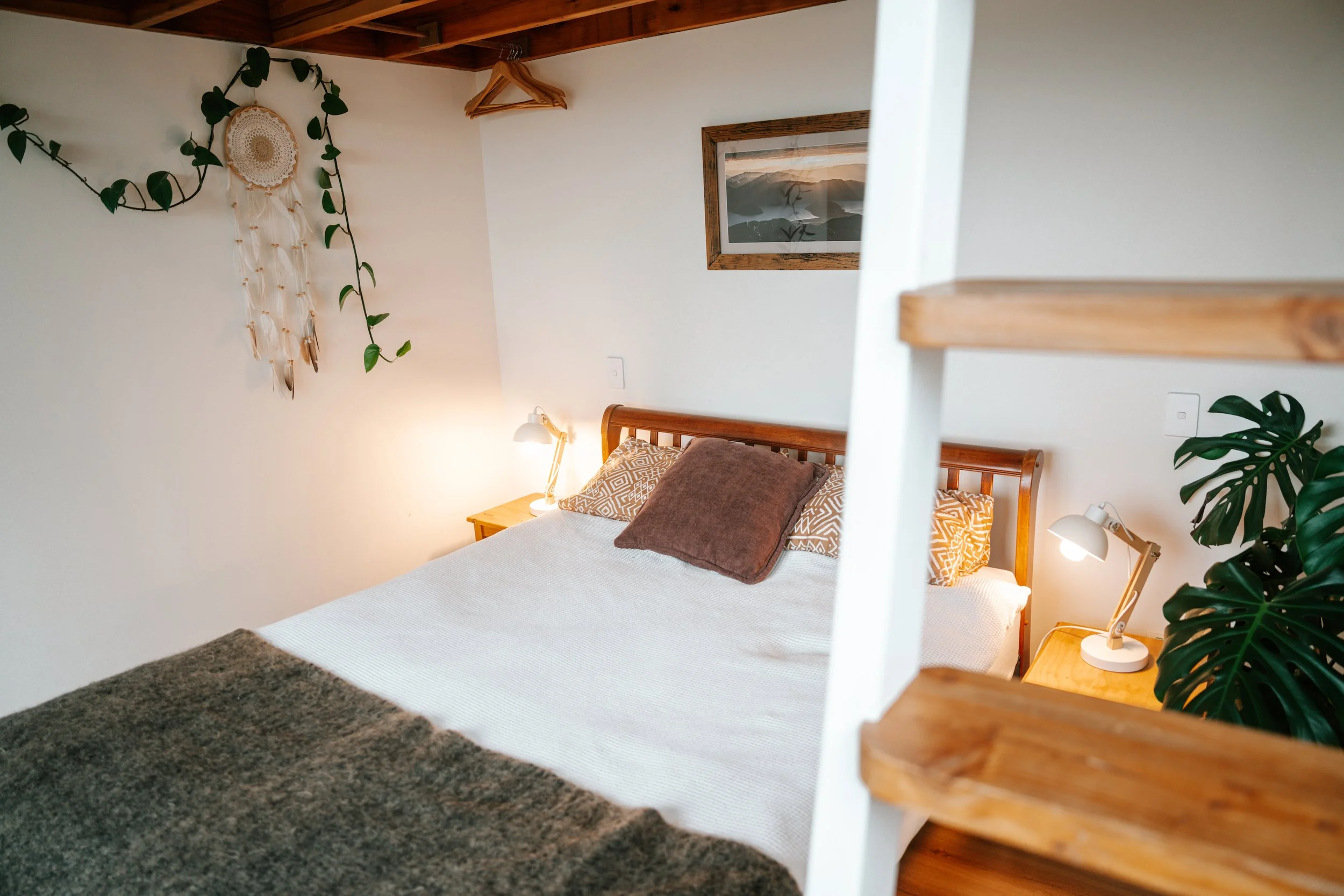 A bedroom with a wooden bed frame, white bedding, brown pillows, wall-mounted lamps, a framed landscape picture, wall hangings, plants, and wooden hangers on a ceiling hook.