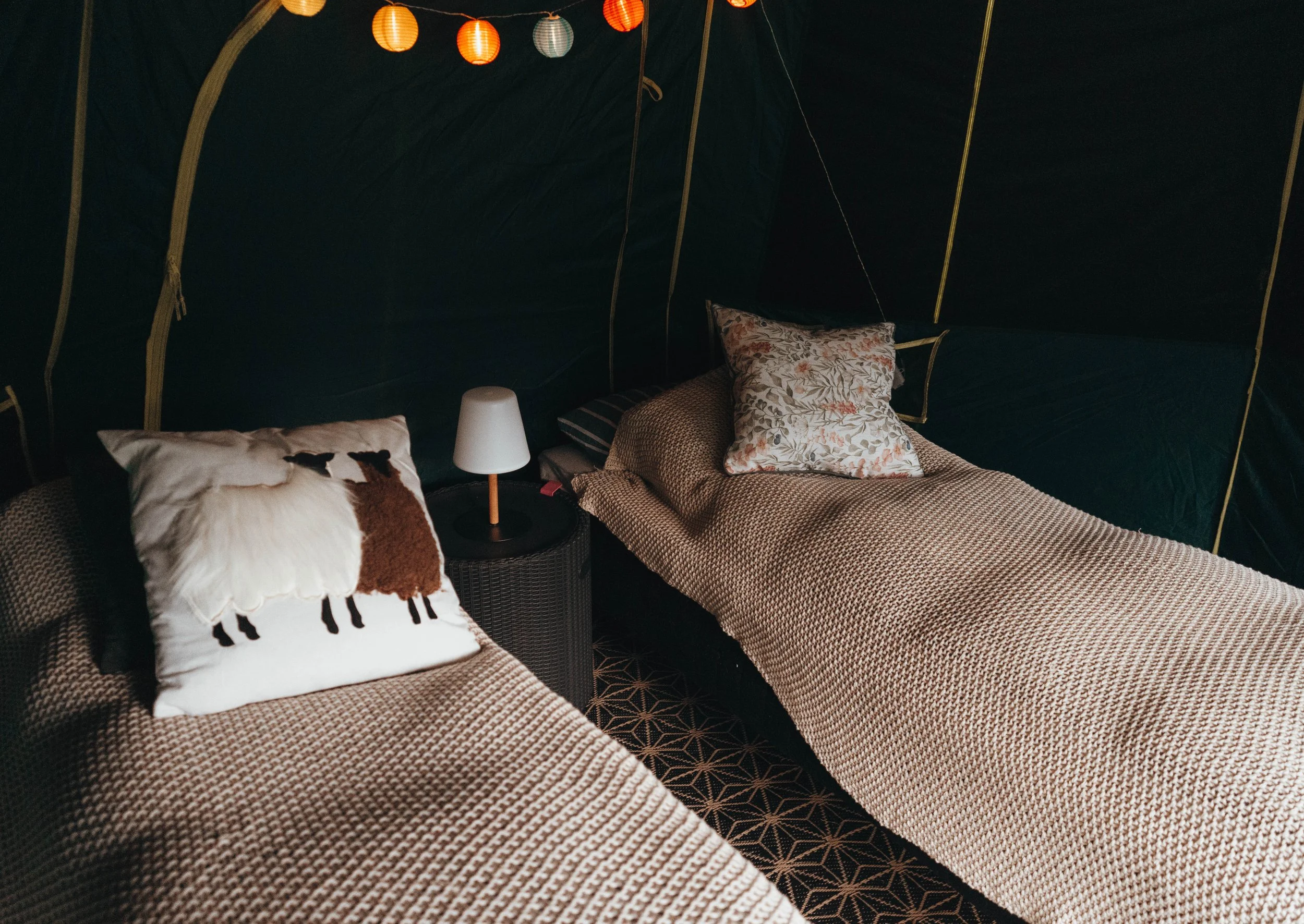 A cozy tent interior decorated with hanging paper lanterns, featuring beds with patterned bedding and pillows, including one with a llama print, and a small white lamp on a round black side table.