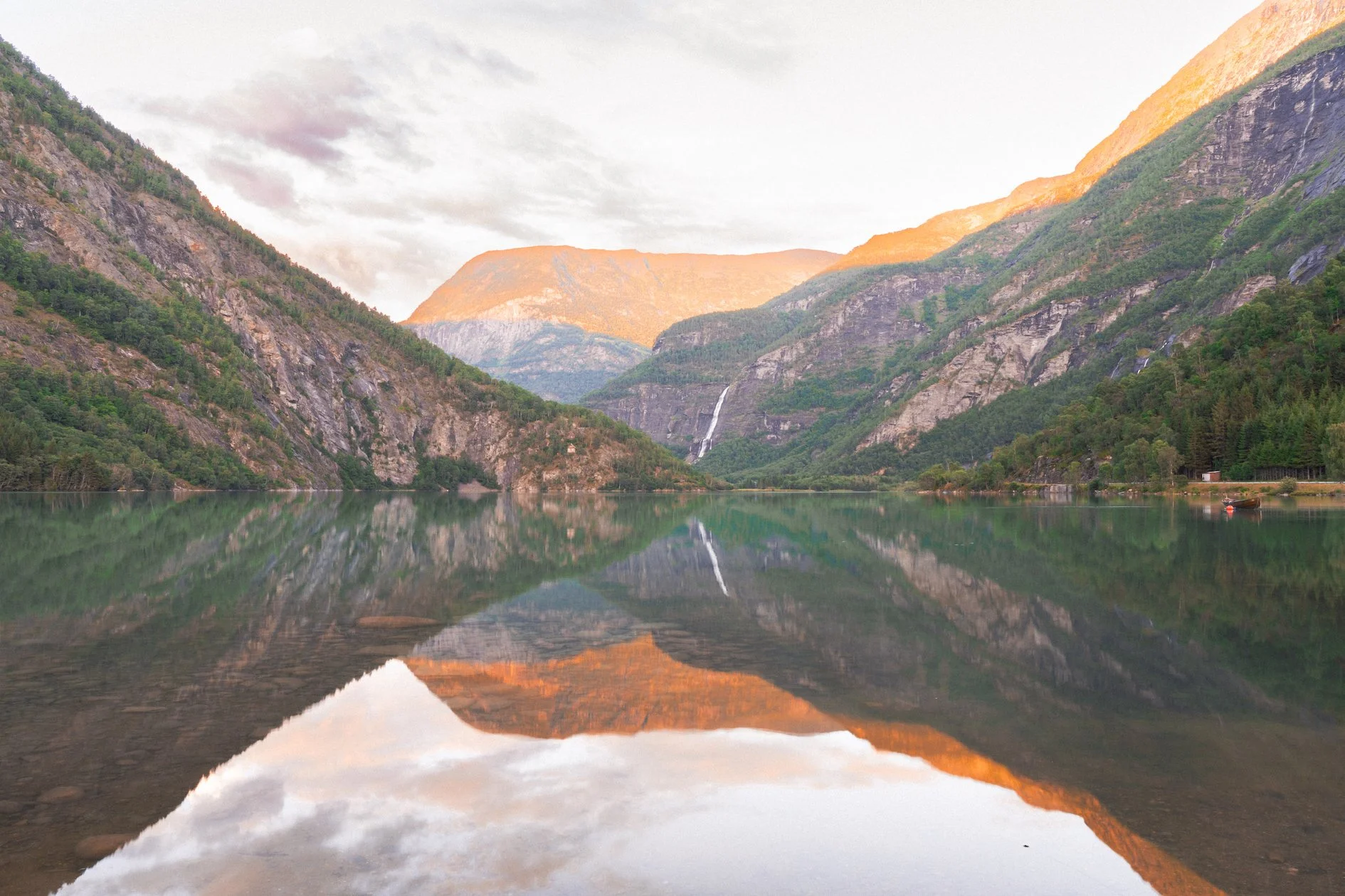 Scenic view of a calm lake surrounded by green mountains with a small boat on the shoreline and a waterfall in the distance, under a partly cloudy sky.