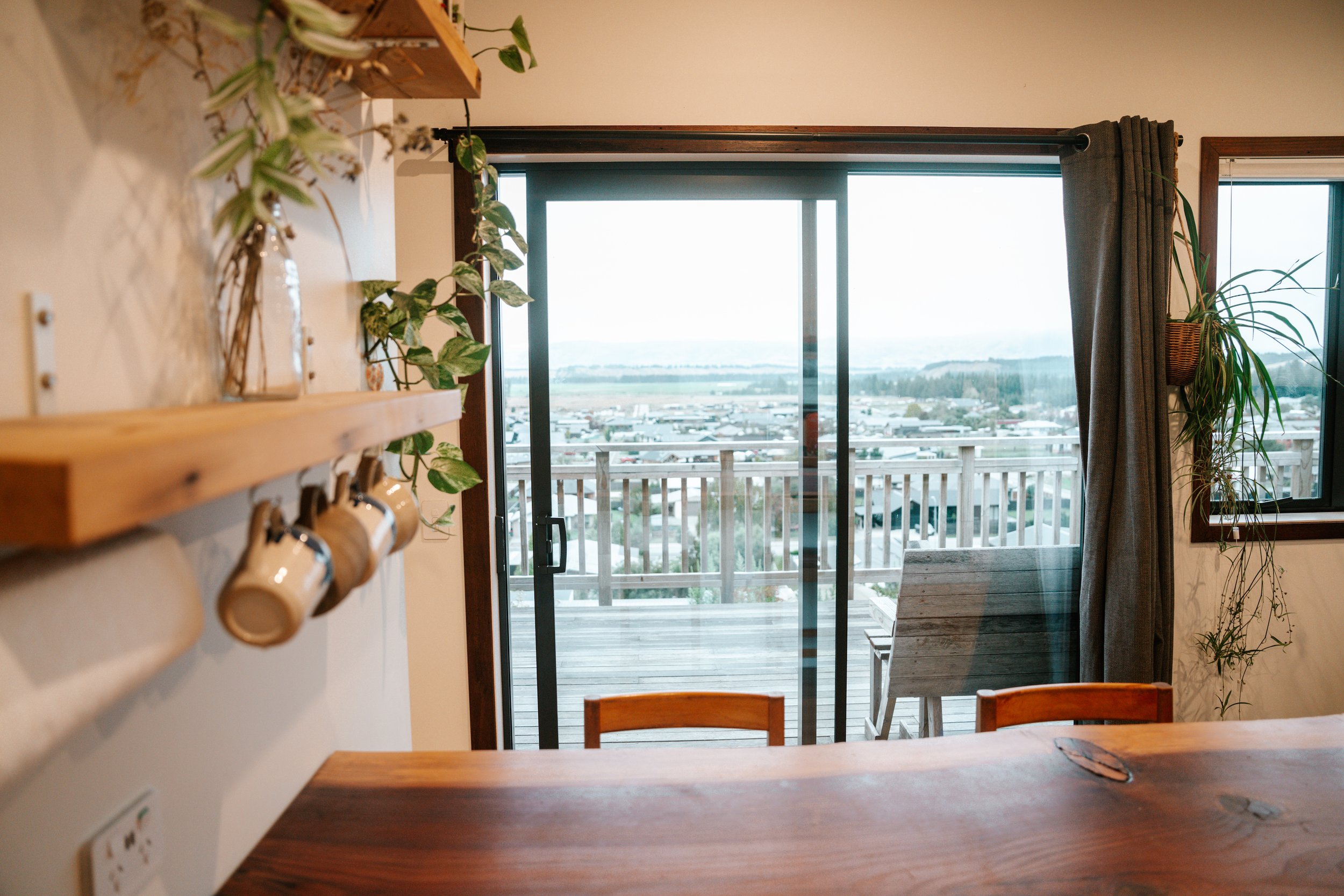 View of a living room with sliding glass doors leading to a balcony, overlooking a suburban landscape. There are houseplants near the door, a curtain on one side, and a wooden table partly visible in the foreground.