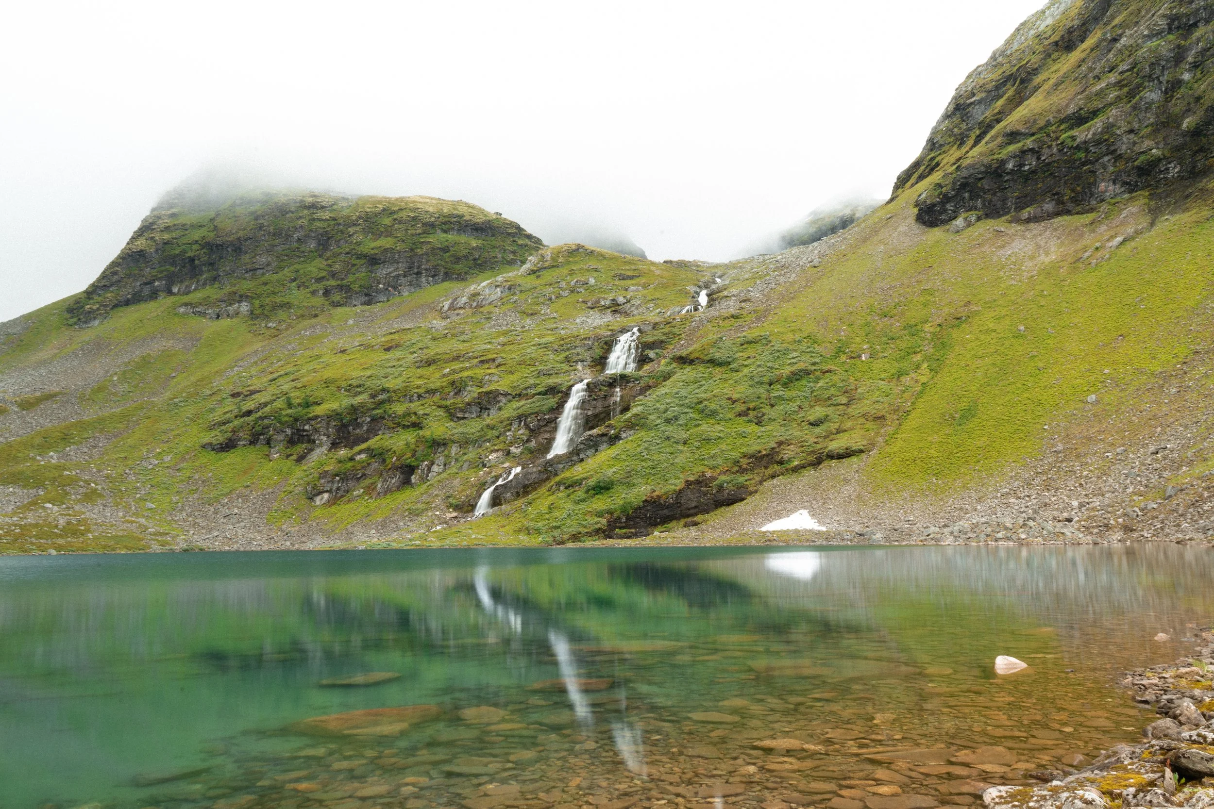Mountain landscape with green slopes, a small waterfall, and a lake in the foreground. Mist covers the top of the mountains.