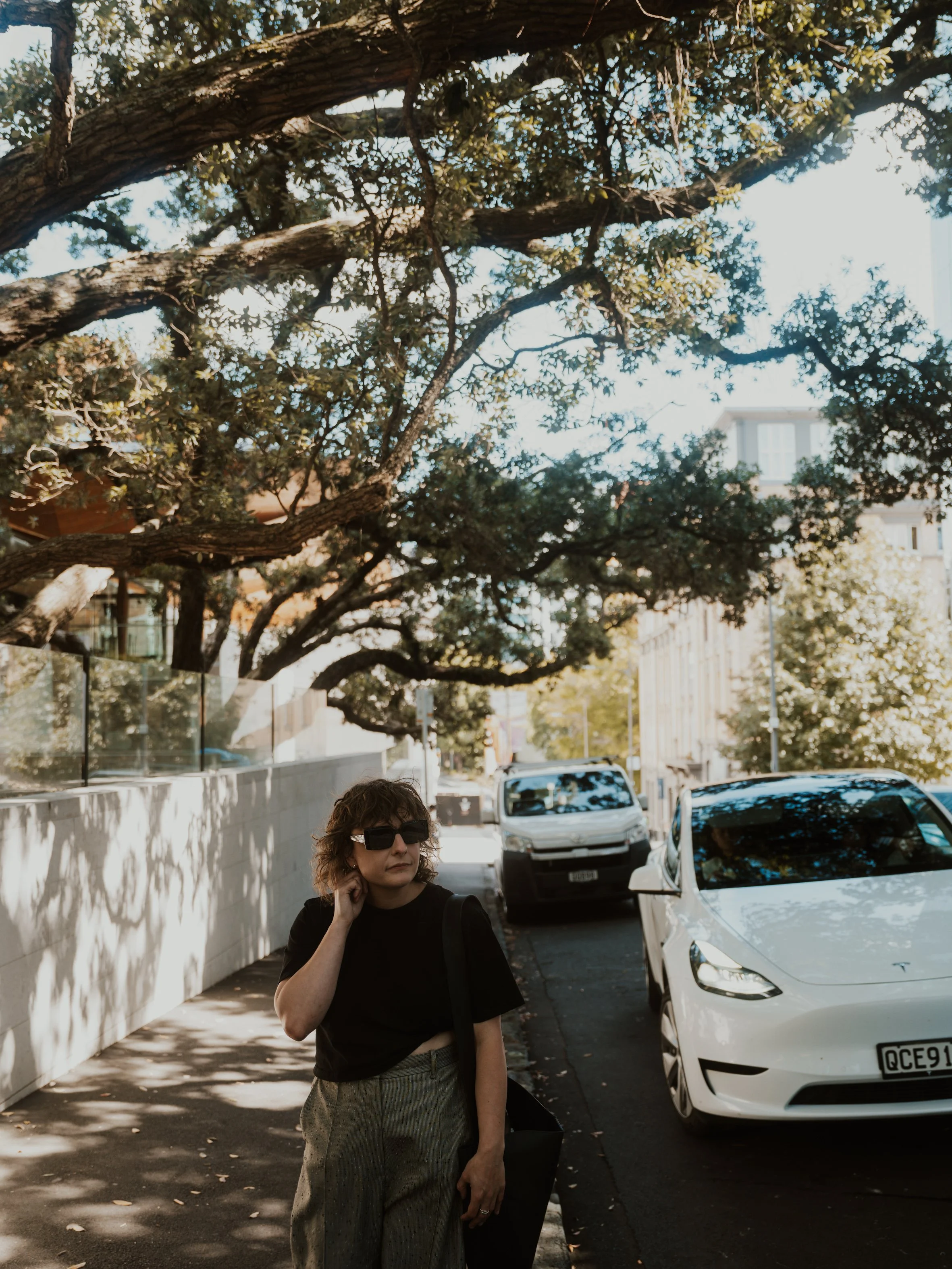 A woman wearing sunglasses, a black top, and gray patterned pants standing on a city street with a white Tesla parked nearby. A large tree with sprawling branches is overhead, and buildings can be seen in the background.