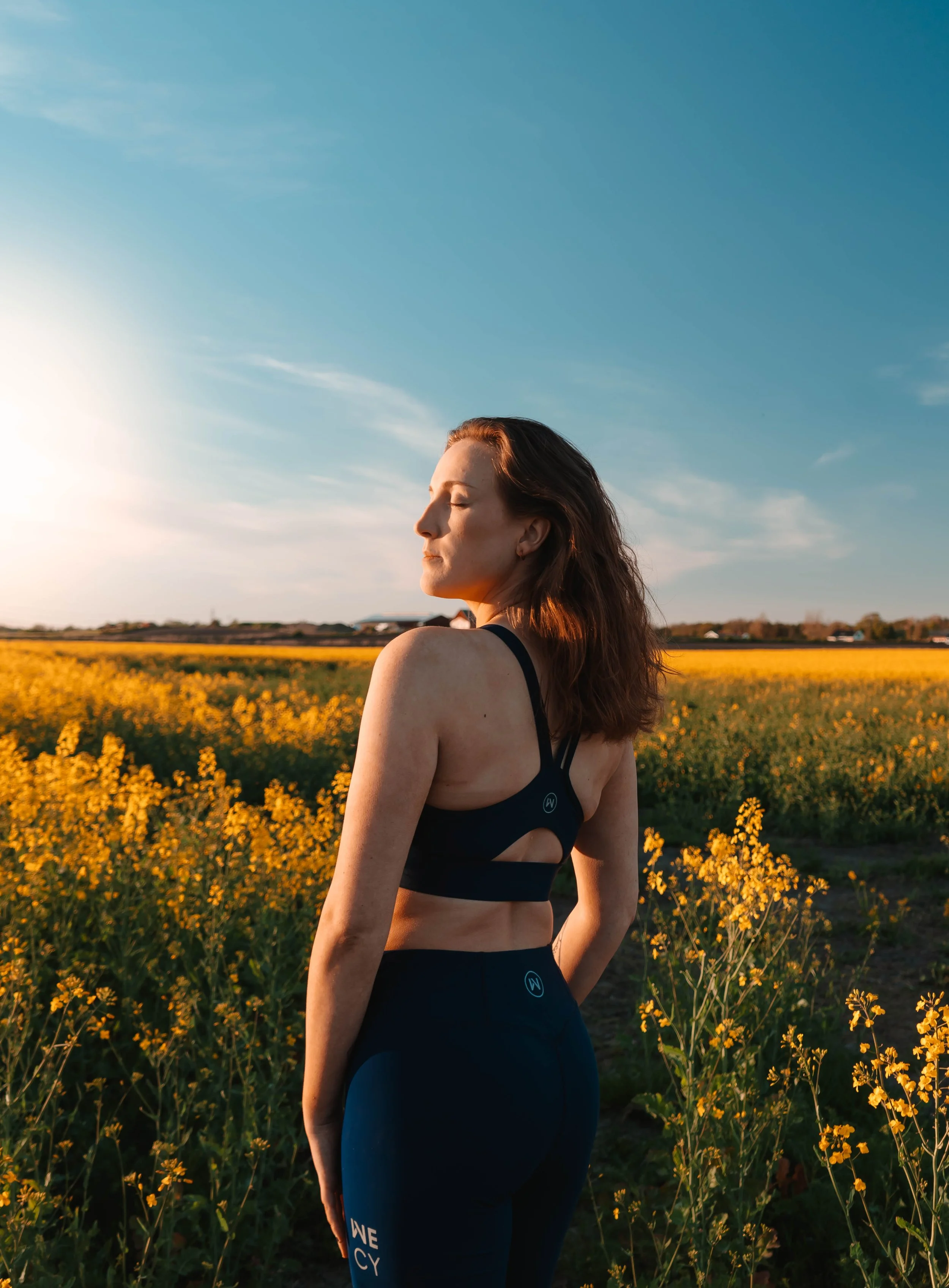 A woman with closed eyes standing in a field of yellow flowers during sunset, wearing a black sports bra and leggings.