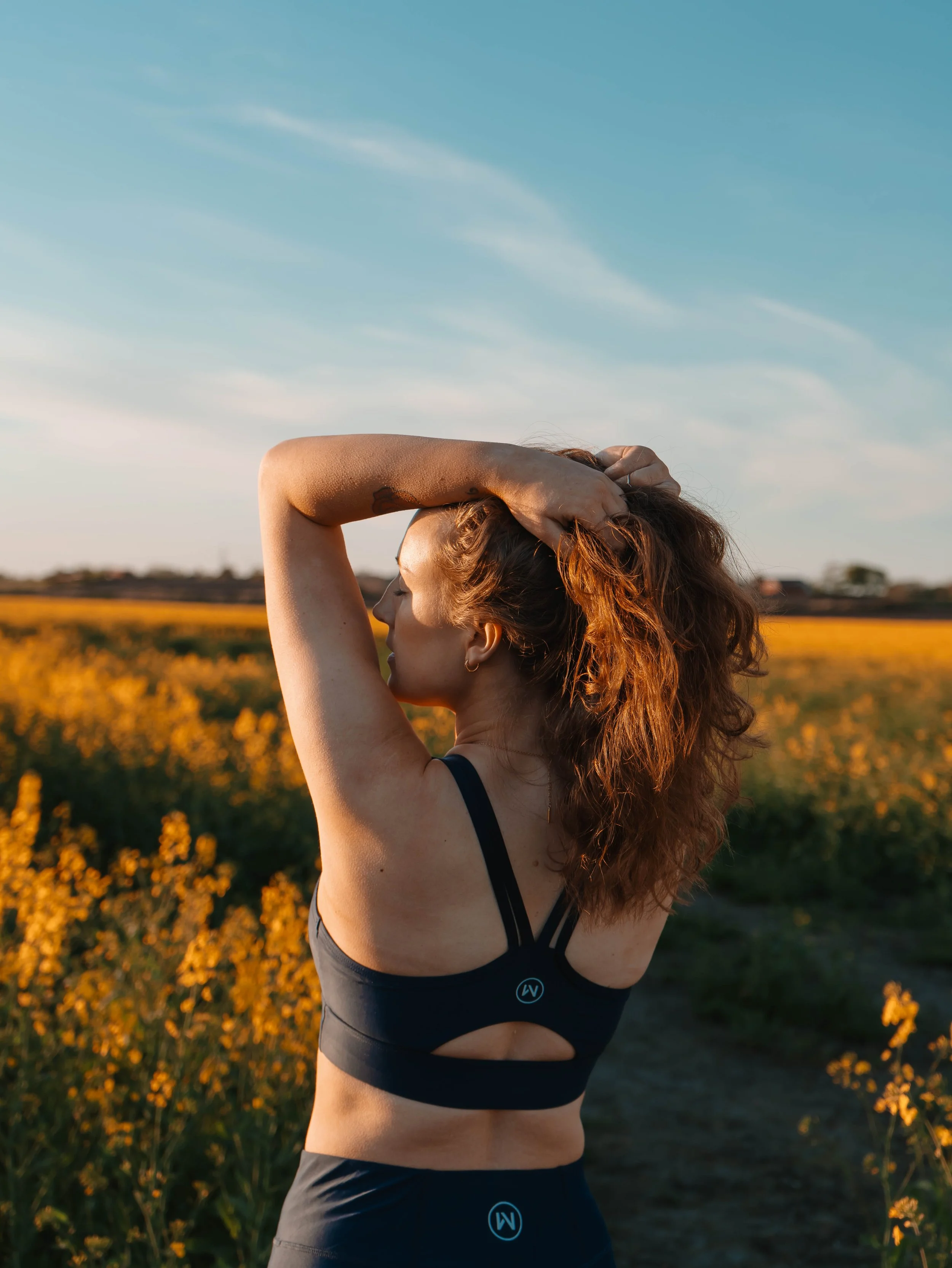 A woman with curly red hair standing outdoors in a field of yellow flowers during sunset, wearing a blue sports bra and matching leggings, with her left hand on her head.