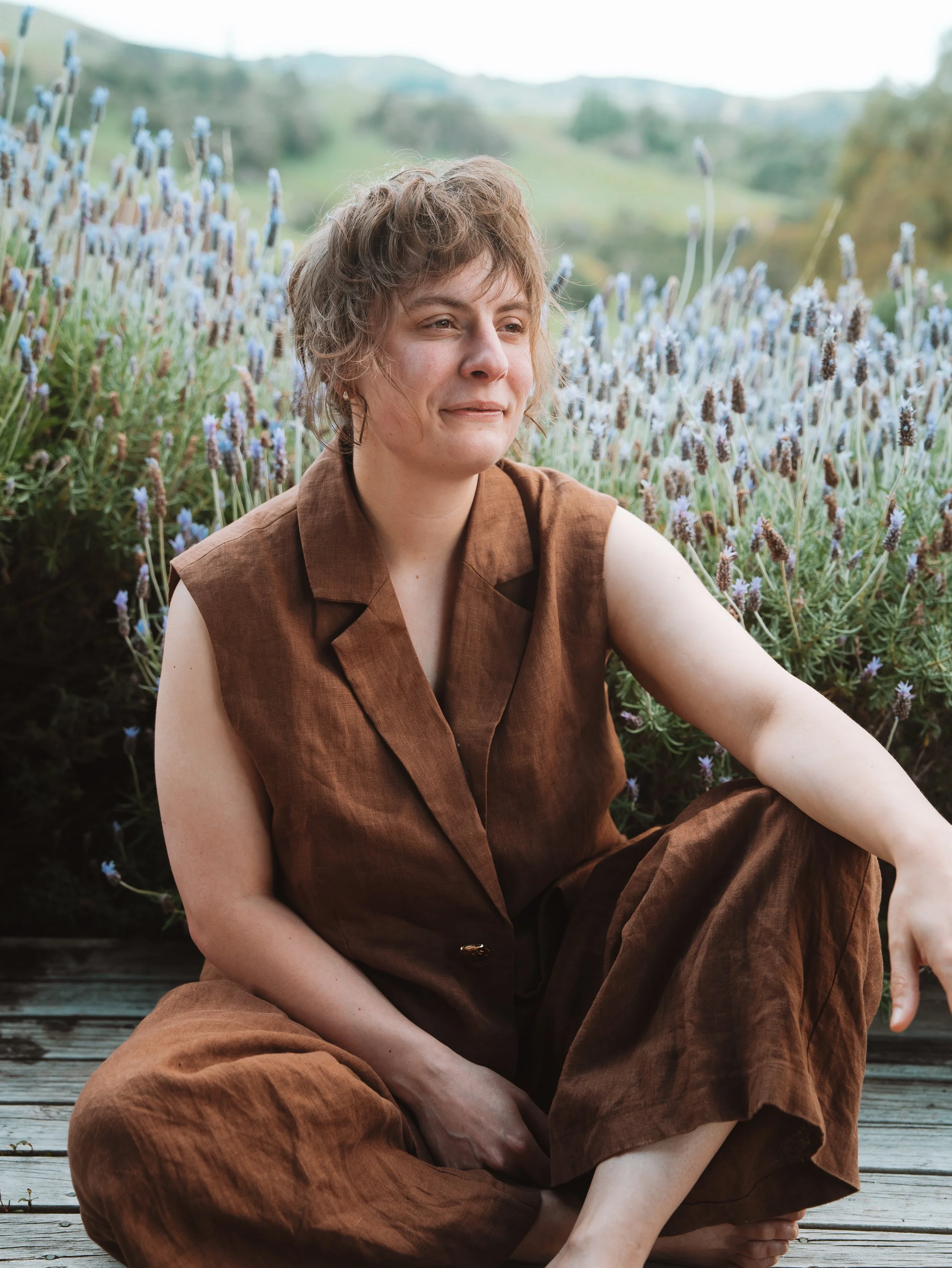 A woman with short, wavy hair sitting outdoors on a wooden surface, surrounded by lavender plants, with a blurred hillside in the background.