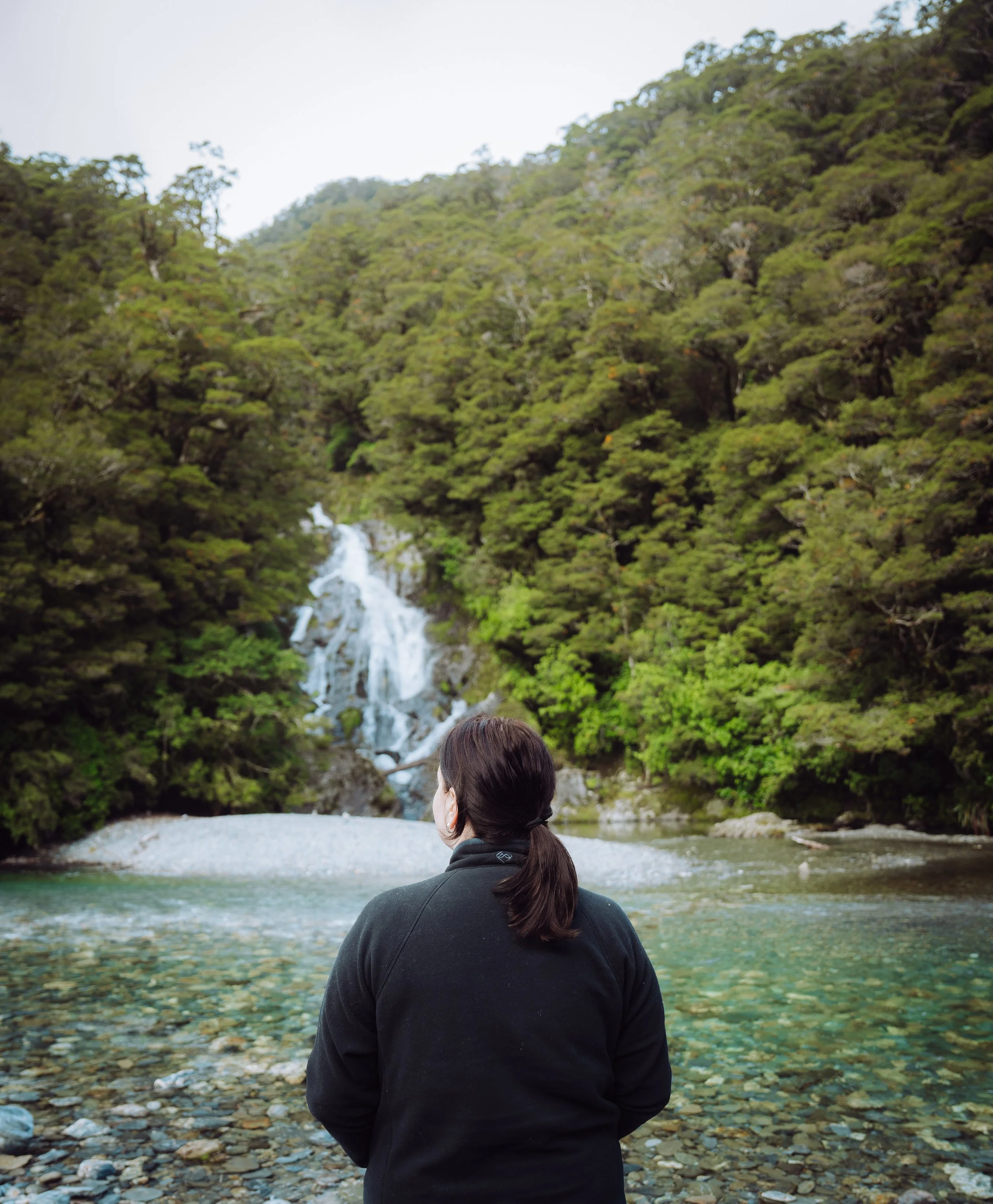 A person with dark hair tied back, wearing a black jacket, standing by a river with a lush green forest and waterfall in the background.