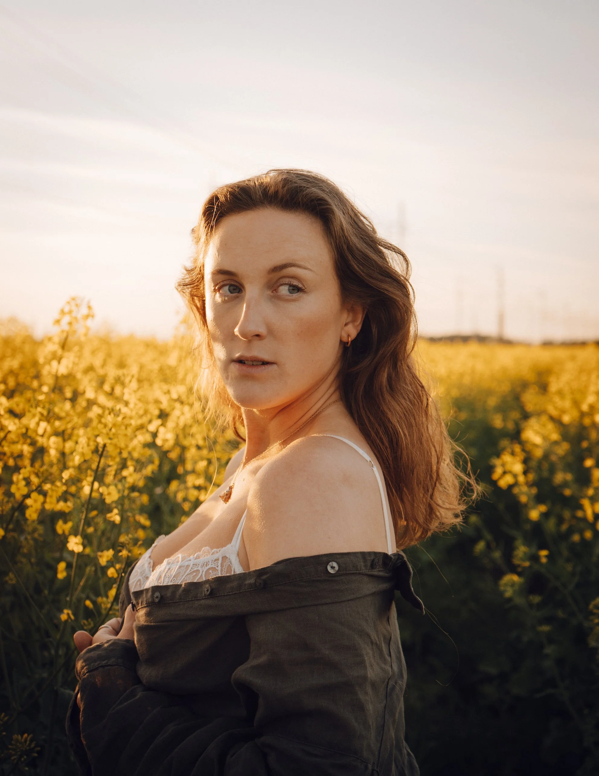 A woman with brown hair looking over her shoulder in a field of yellow flowers during sunset.