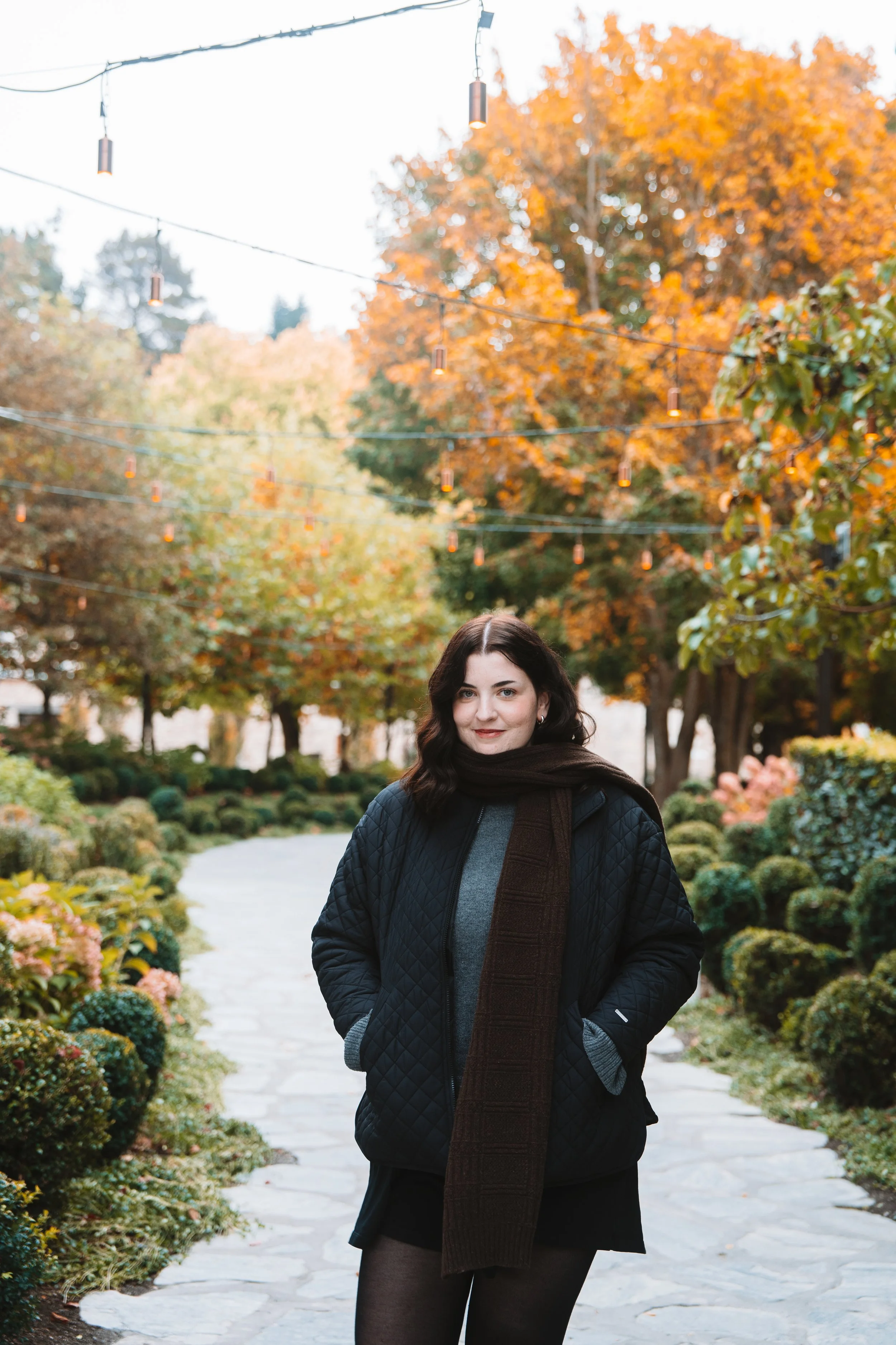 A woman standing on a stone pathway in a garden with orange autumn leaves, hanging string lights, and trees in the background.