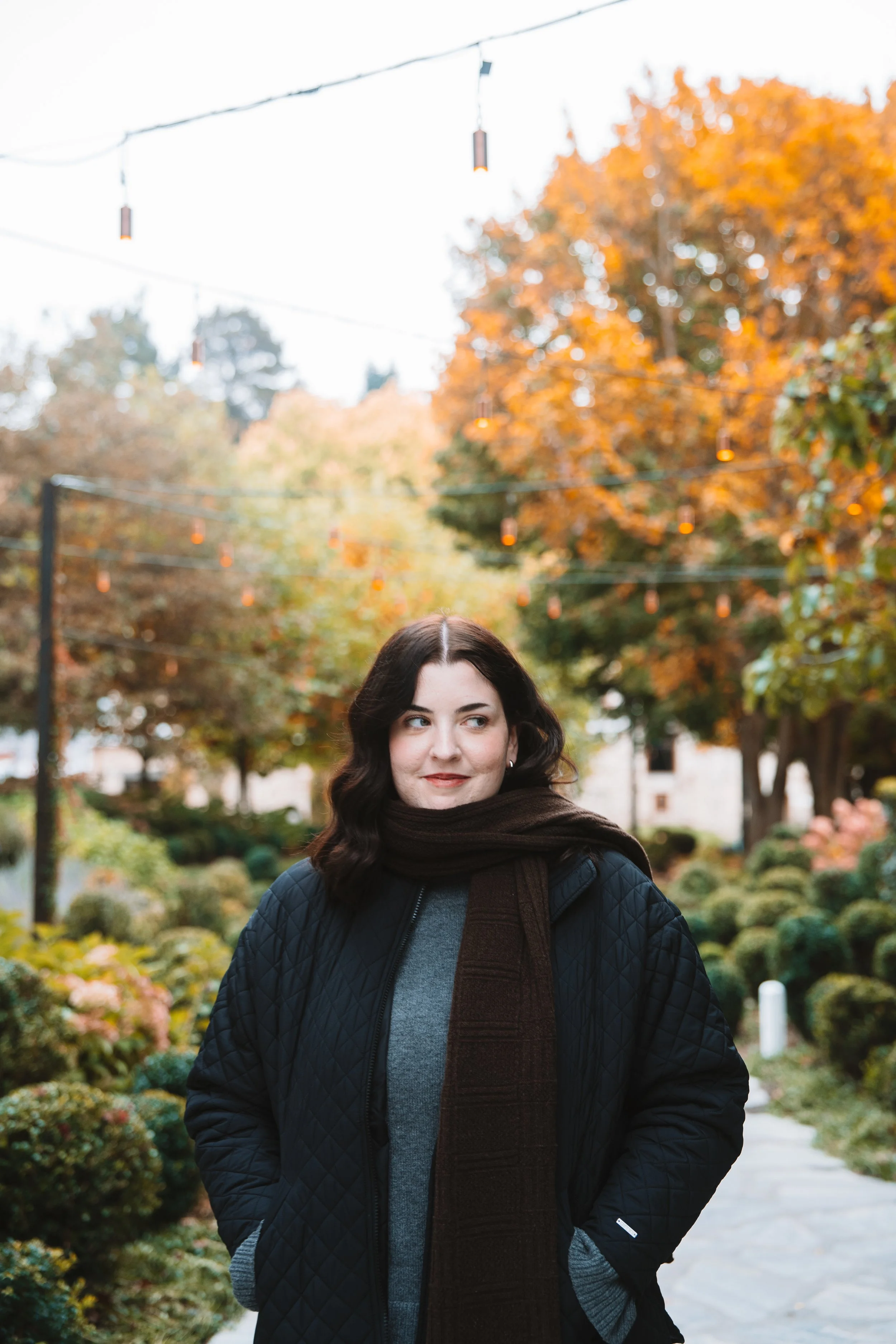 A woman with dark wavy hair and light skin standing outdoors in fall foliage, wearing a black quilted jacket, gray sweater, and a brown scarf, looking to her left with a slight smile.