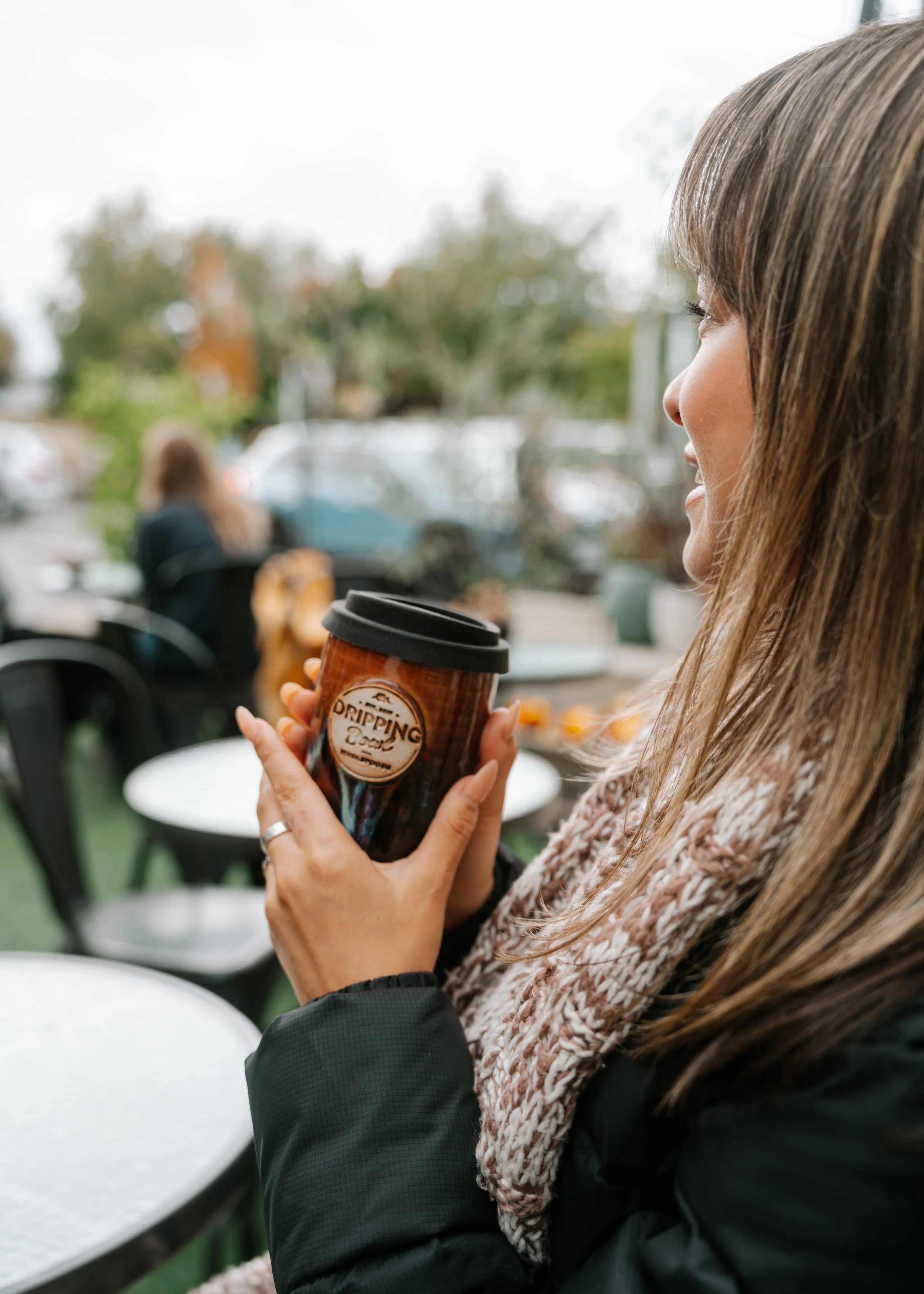 A woman with long brown hair, wearing a black jacket and a beige scarf, holding a coffee cup and smiling.