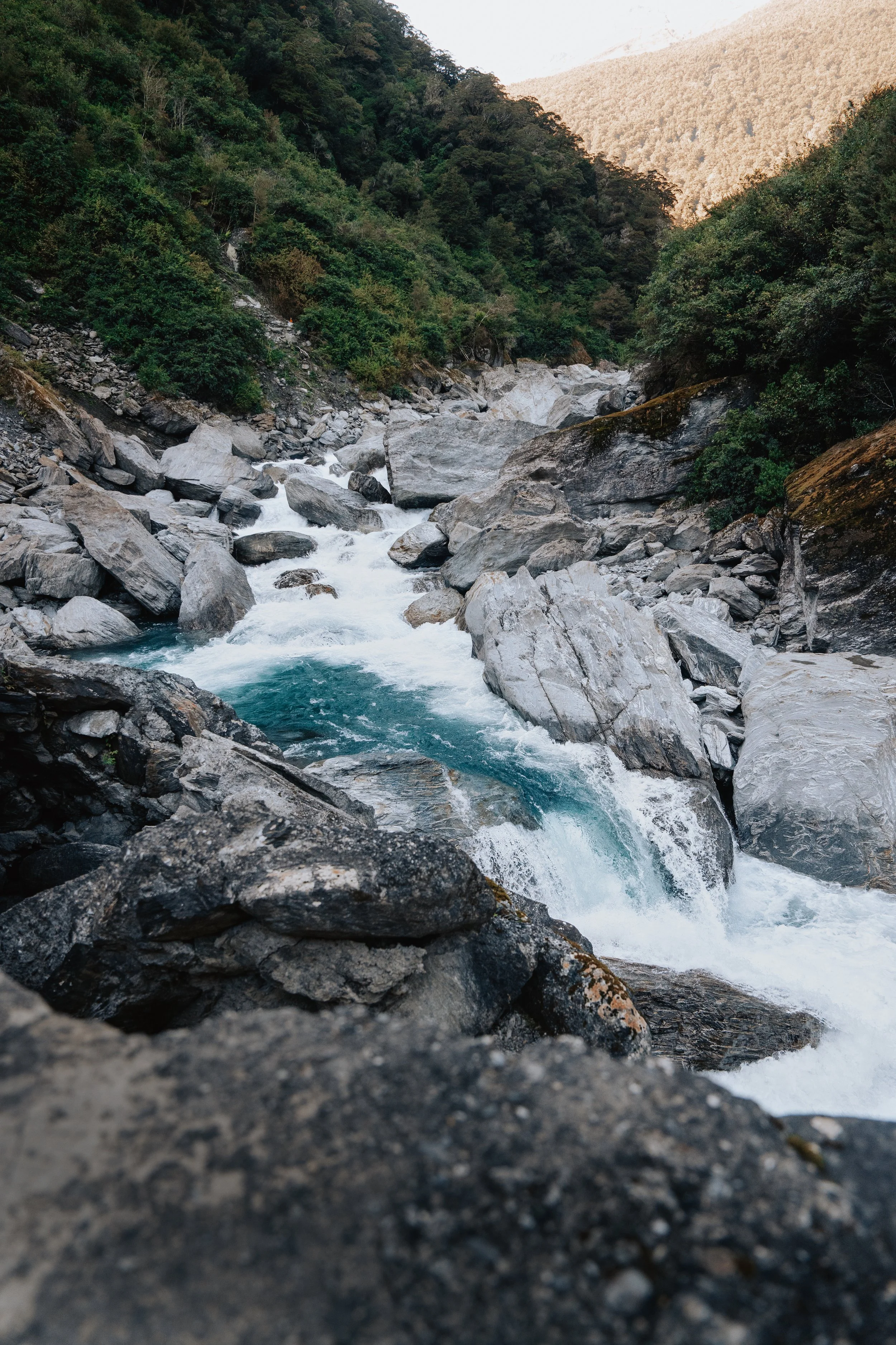 Flowing river through rocky terrain surrounded by green forested hills with in the background mountains.