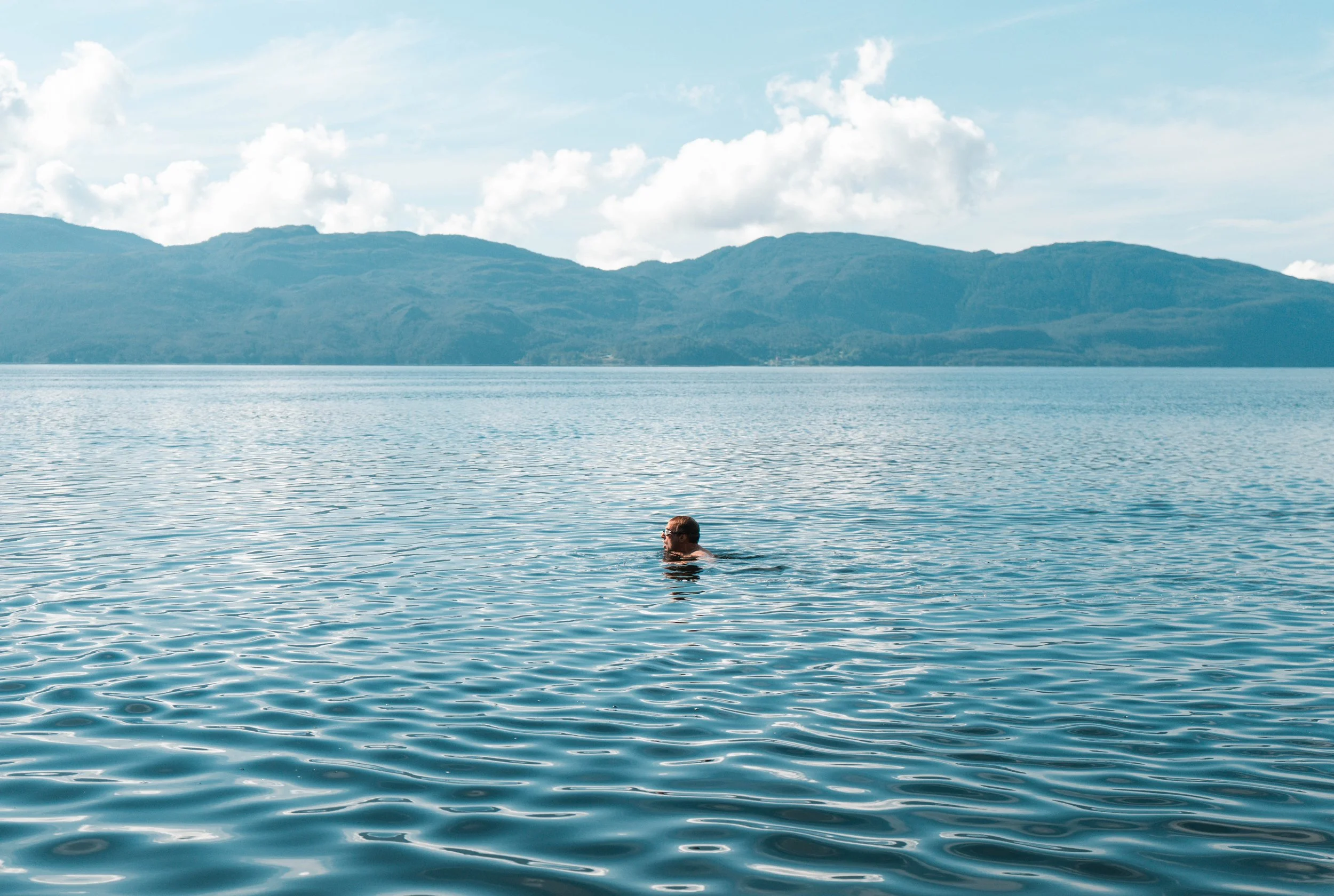 A person swimming in a body of water with mountain ranges and a partly cloudy sky in the background.