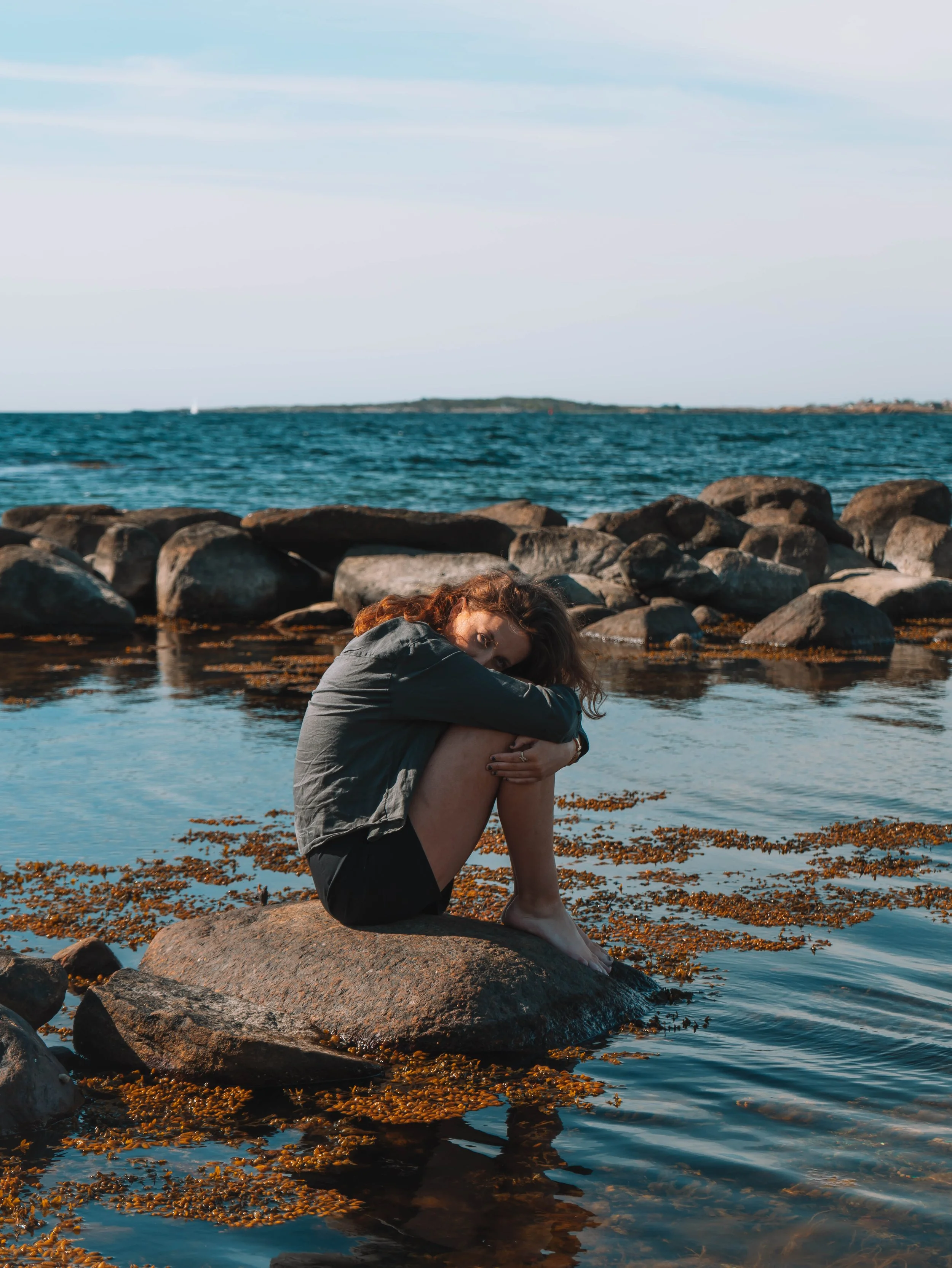 Woman sitting on a rock by the water with rocks in the background, wearing a gray jacket and black shorts, with curly red hair, during sunset or late afternoon at a beach.