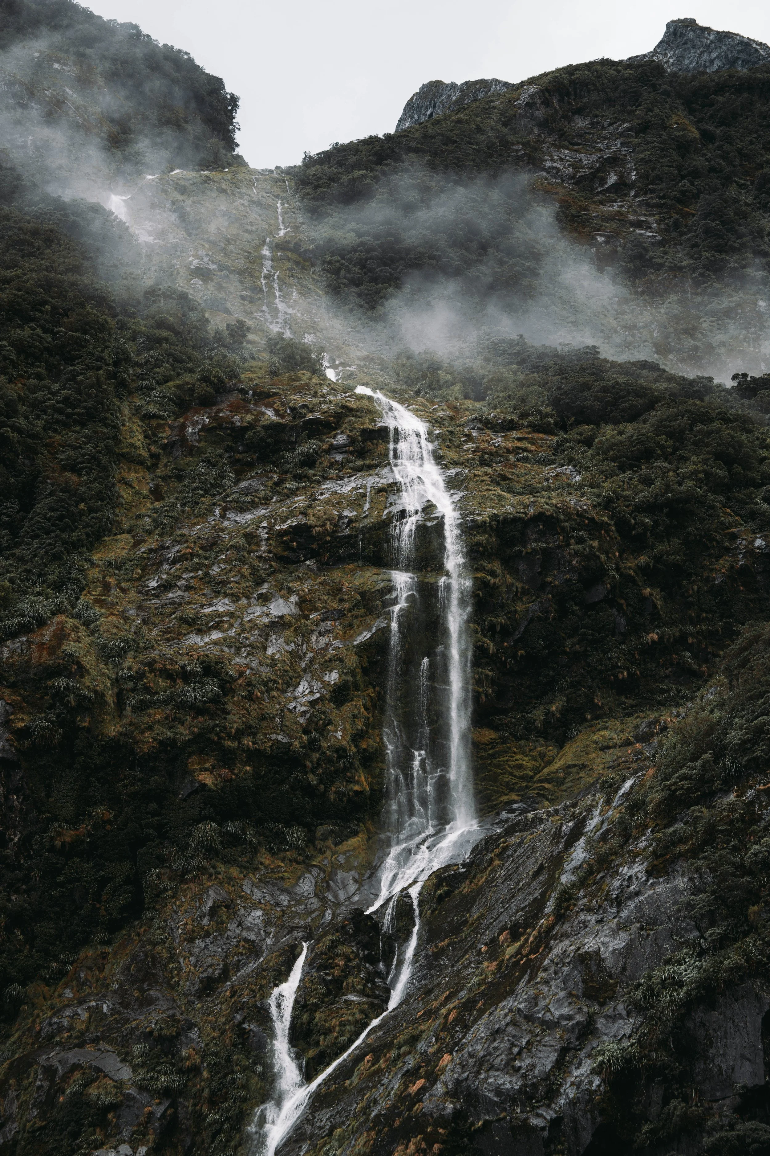 A tall mountain with a waterfall cascading down its rocky, moss-covered slope, with mist and clouds surrounding the peak.