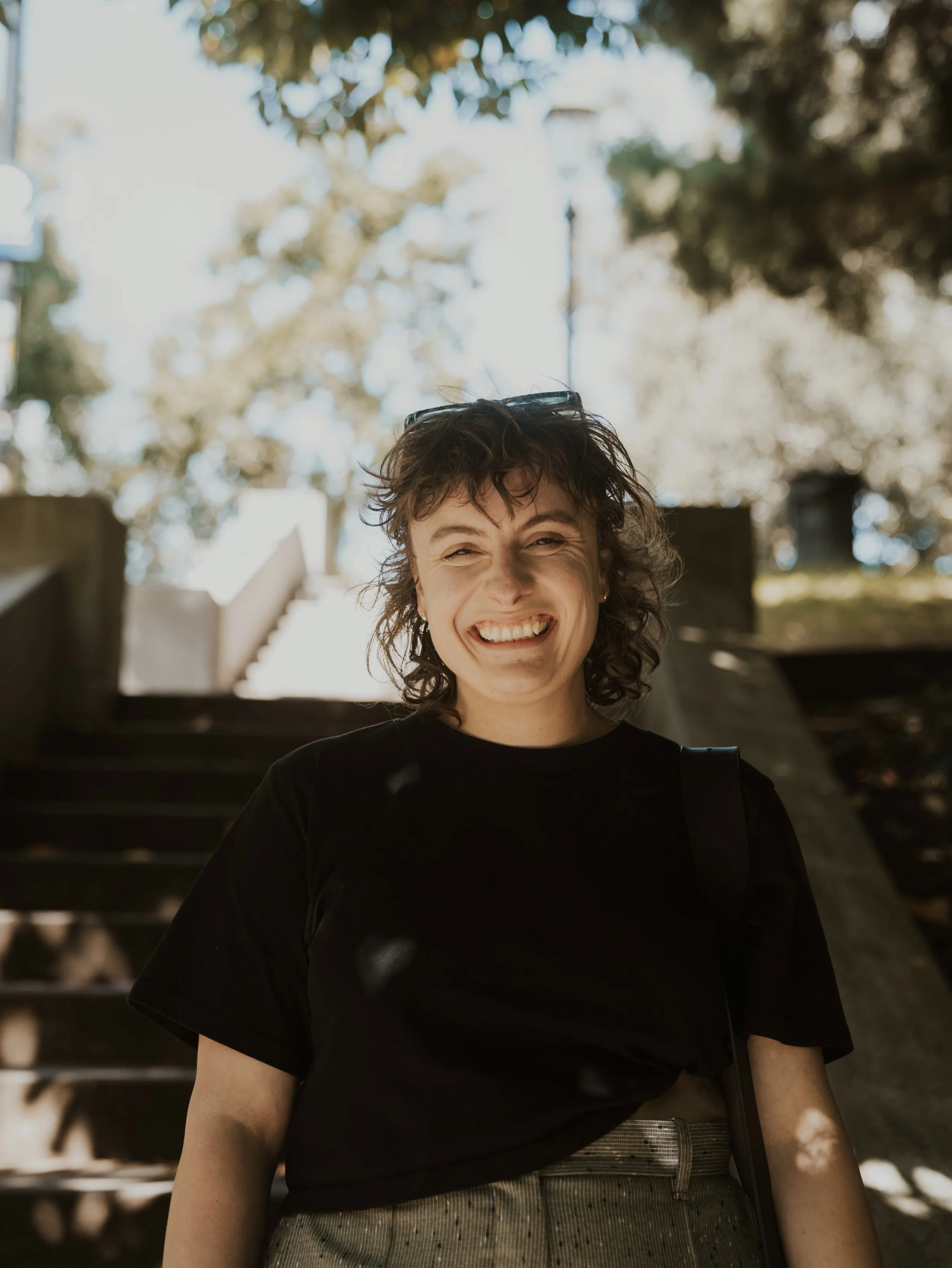 Smiling woman with curly hair wearing a black shirt and beige pants standing outdoors on stairs with trees in the background.