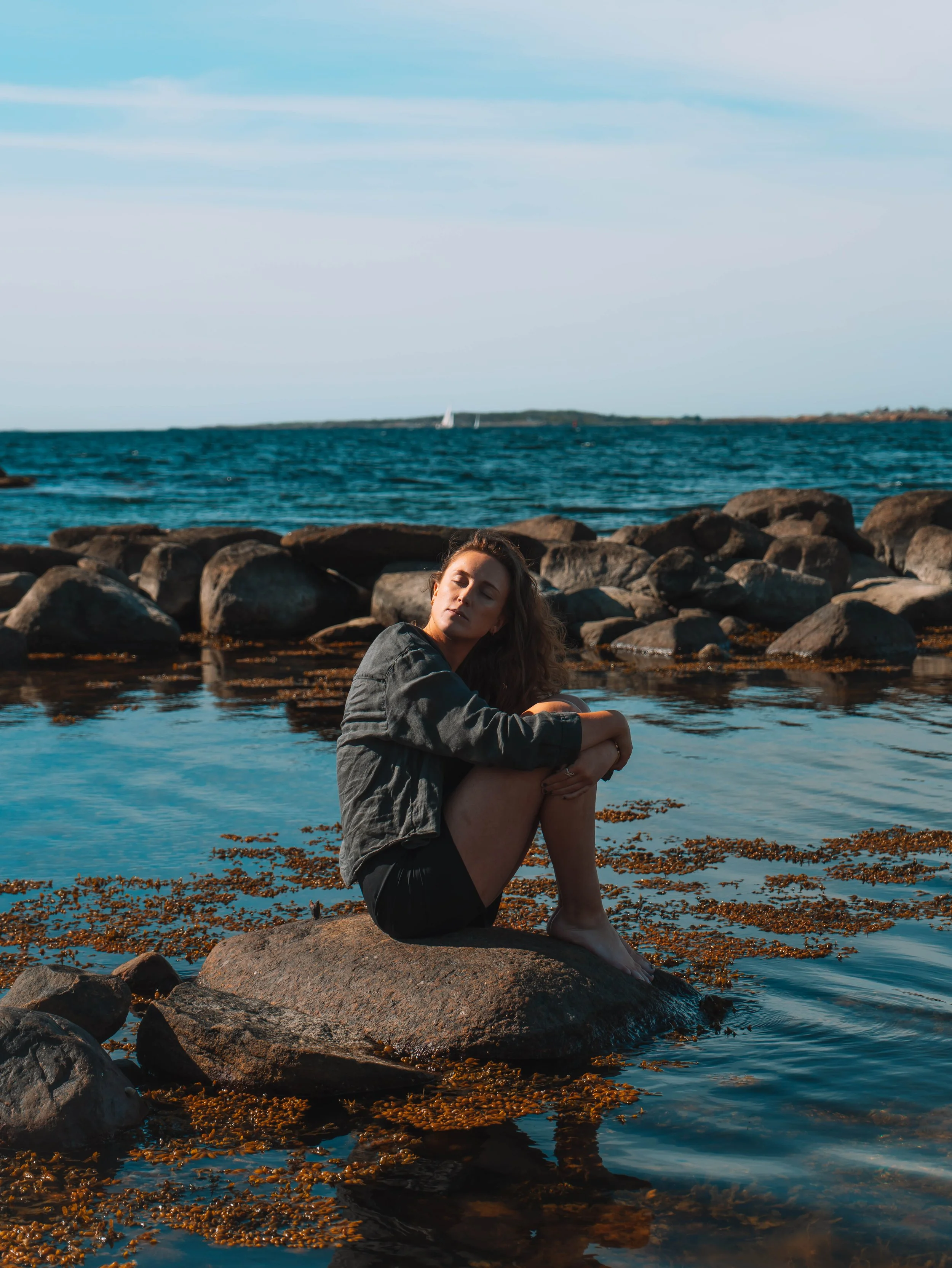 A young woman sitting on a large rock at the edge of the water, with a rocky shoreline behind her and ocean waves. She has long curly hair and is wearing a jacket. The background features a calm sea with a sailboat and a distant shoreline under a par