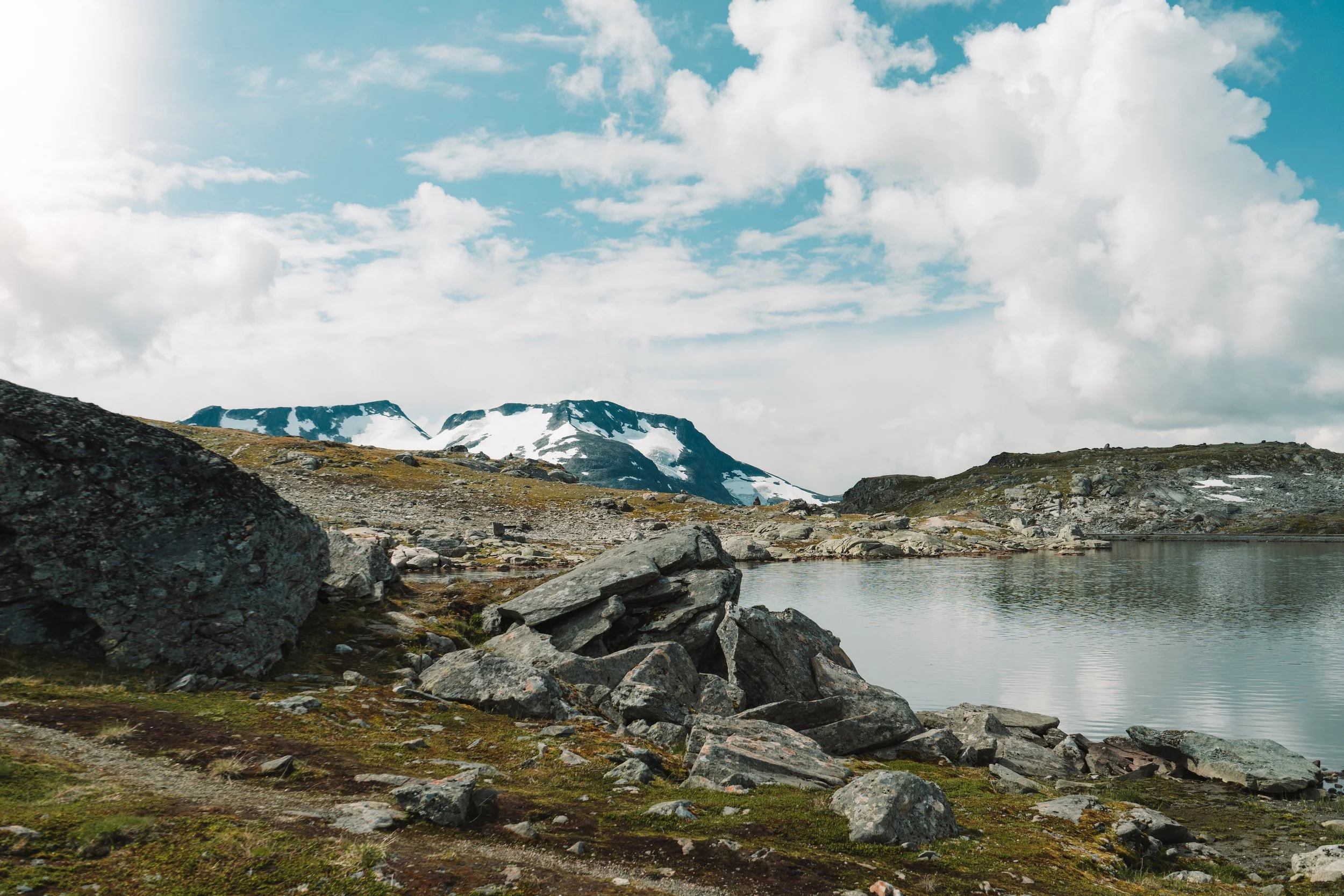 Mountain landscape with snow-capped peaks, rocky foreground, a small lake, and a partly cloudy sky.