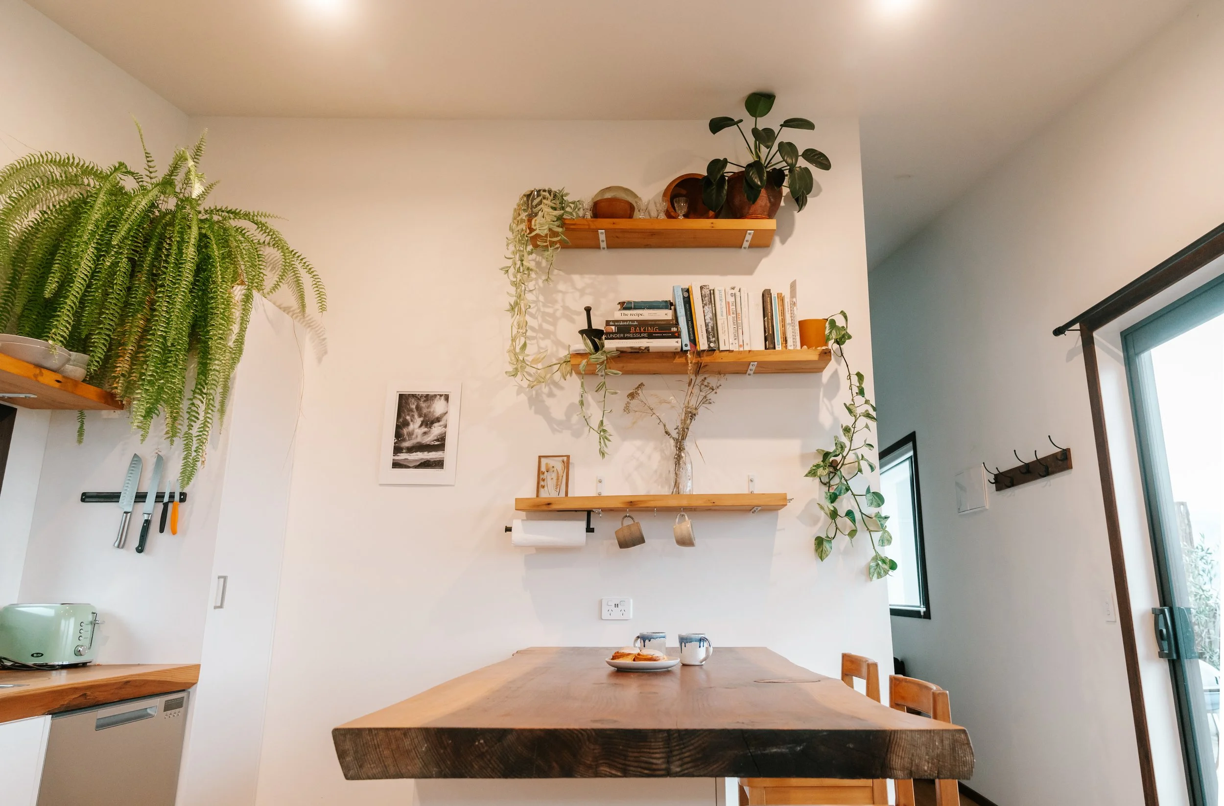 A kitchen with white walls decorated with wooden shelves holding plants, books, and decorative items. There is a large green fern on the left in a pot, a smaller plant on the right, a framed black and white photo, and utensils hanging on the wall. A 