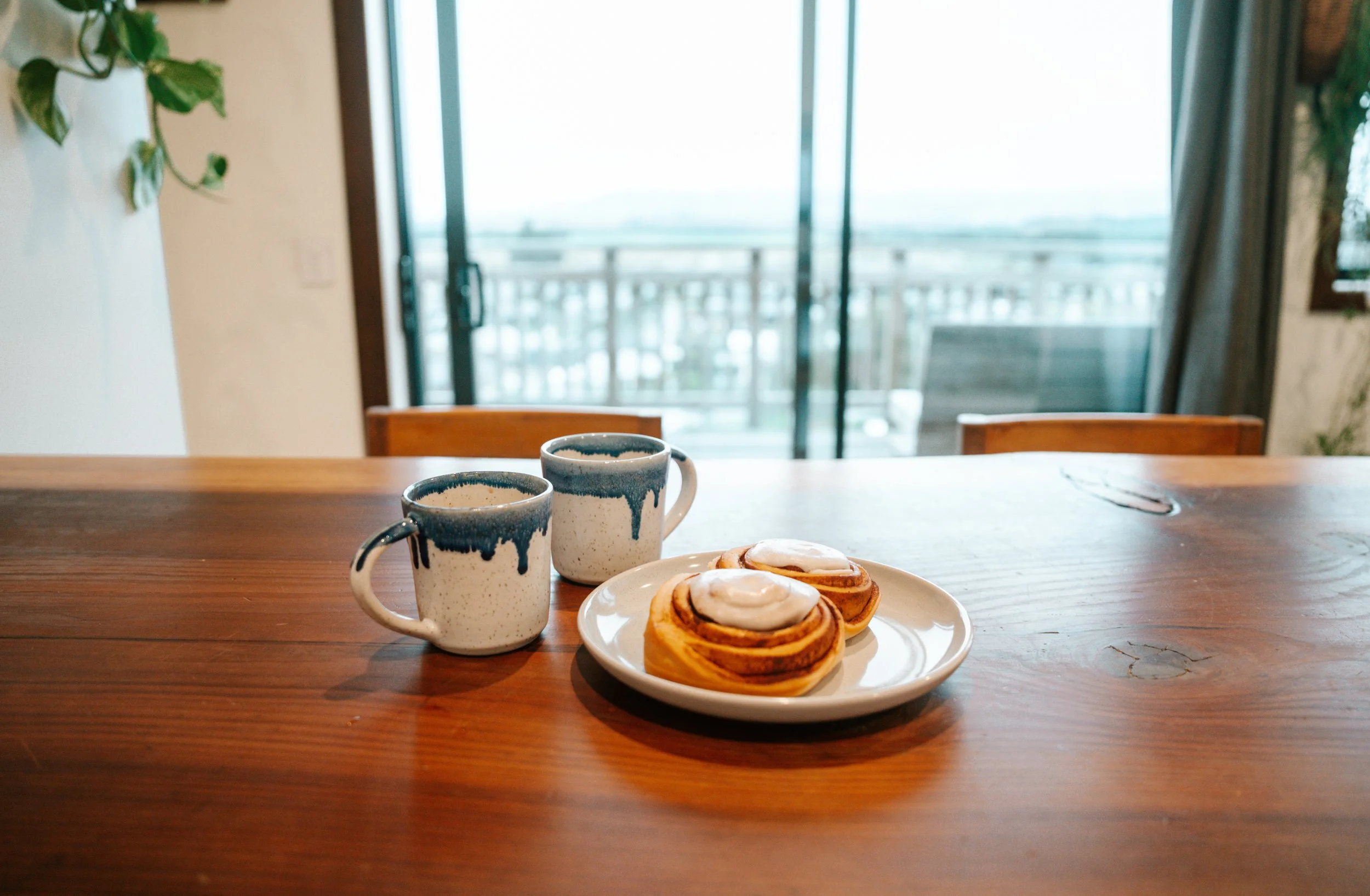 Two ceramic mugs with blue drip glaze on a wooden table next to a plate with three cinnamon rolls topped with icing, against a background of a bright sliding glass door and a balcony.