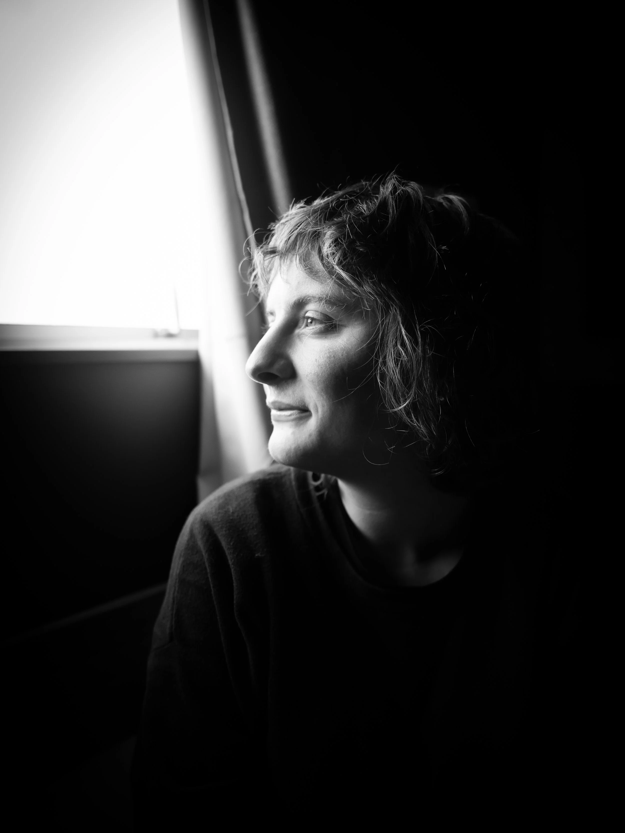 A black and white close-up photo of a young person with casual hair staring out a window, with high contrast lighting highlighting their face against a dark background.
