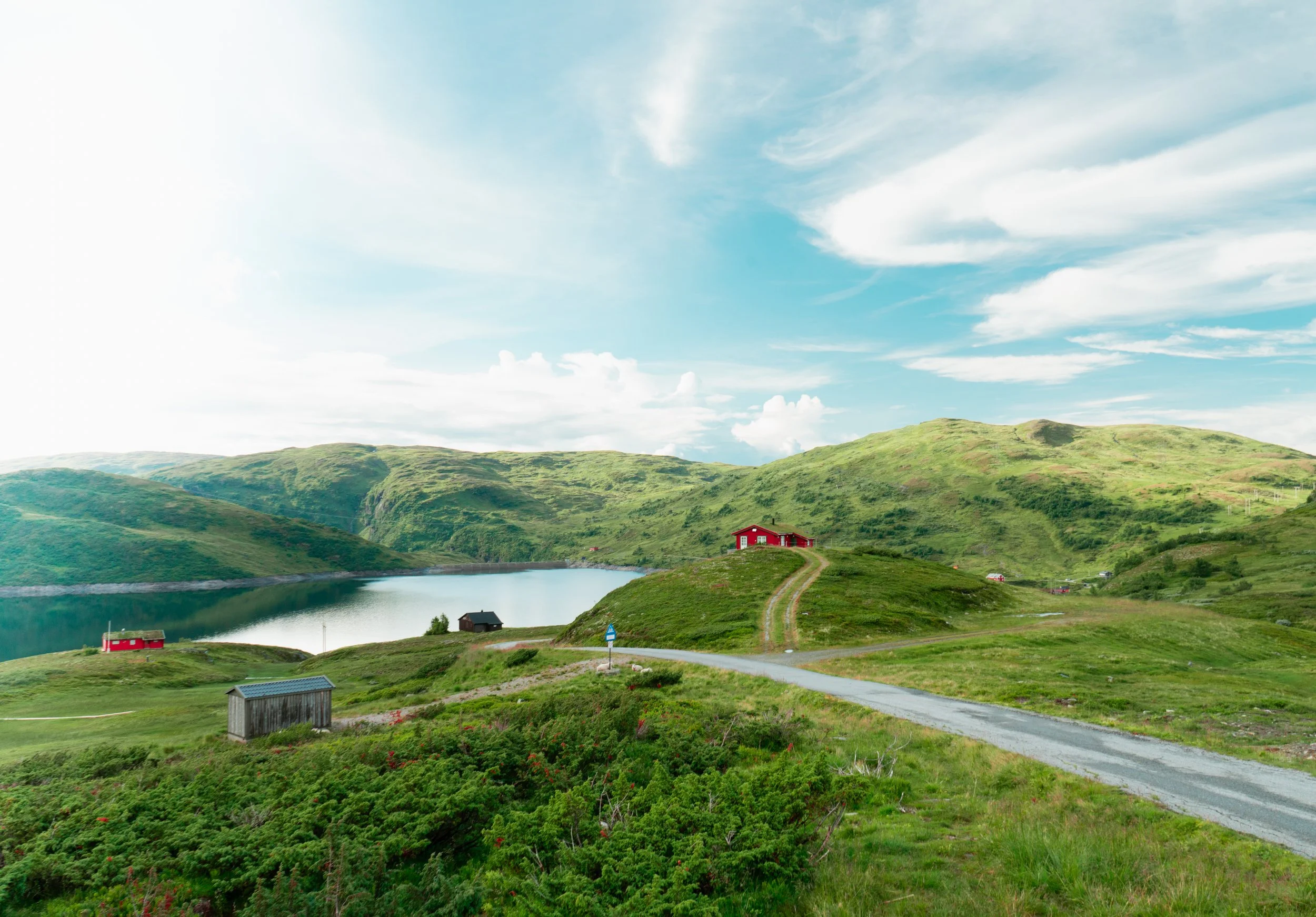 Scenic landscape of a hilly area with green grass, houses, and a lake, under a partly cloudy sky.
