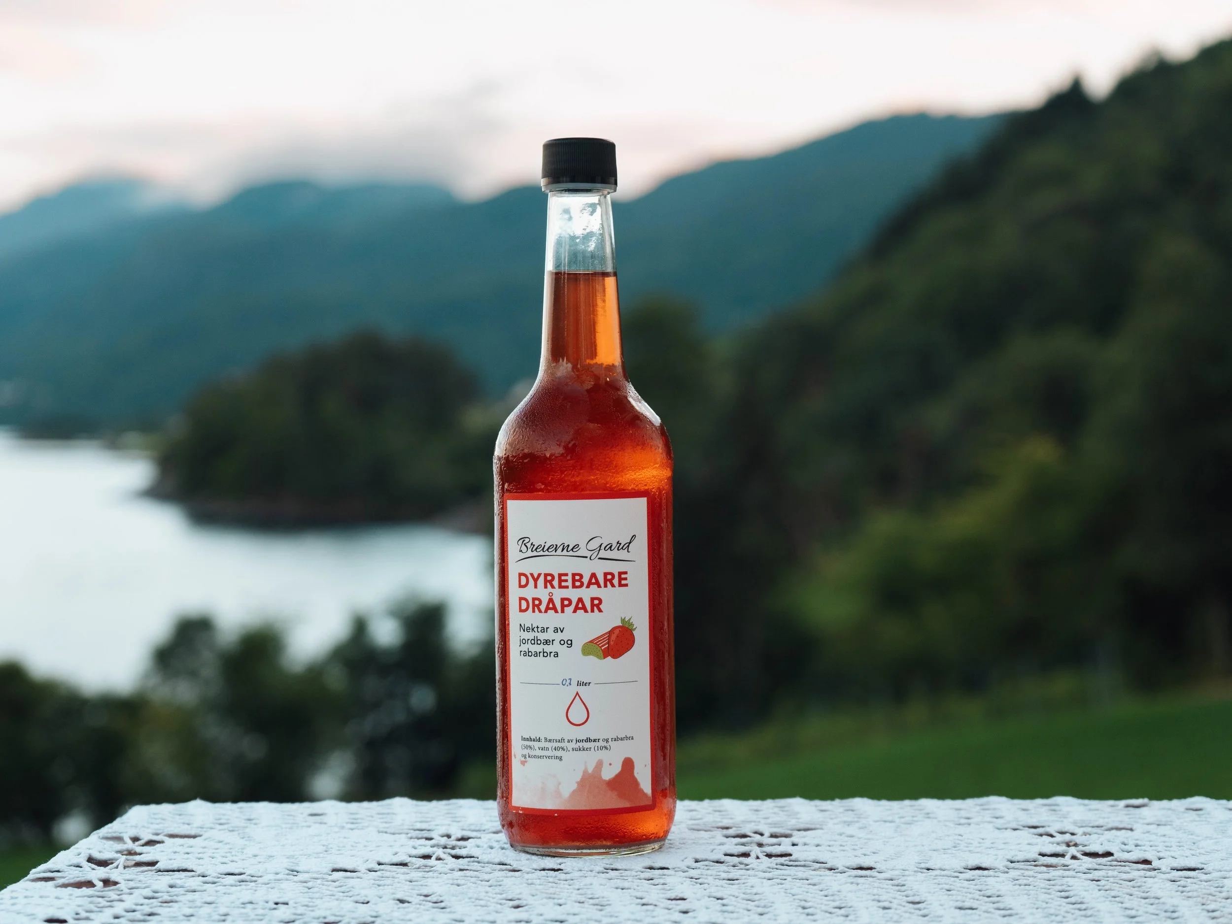 A glass bottle filled with red strawberry and rhubarb juice, placed on a lace tablecloth with a scenic mountain and lake background during daytime.