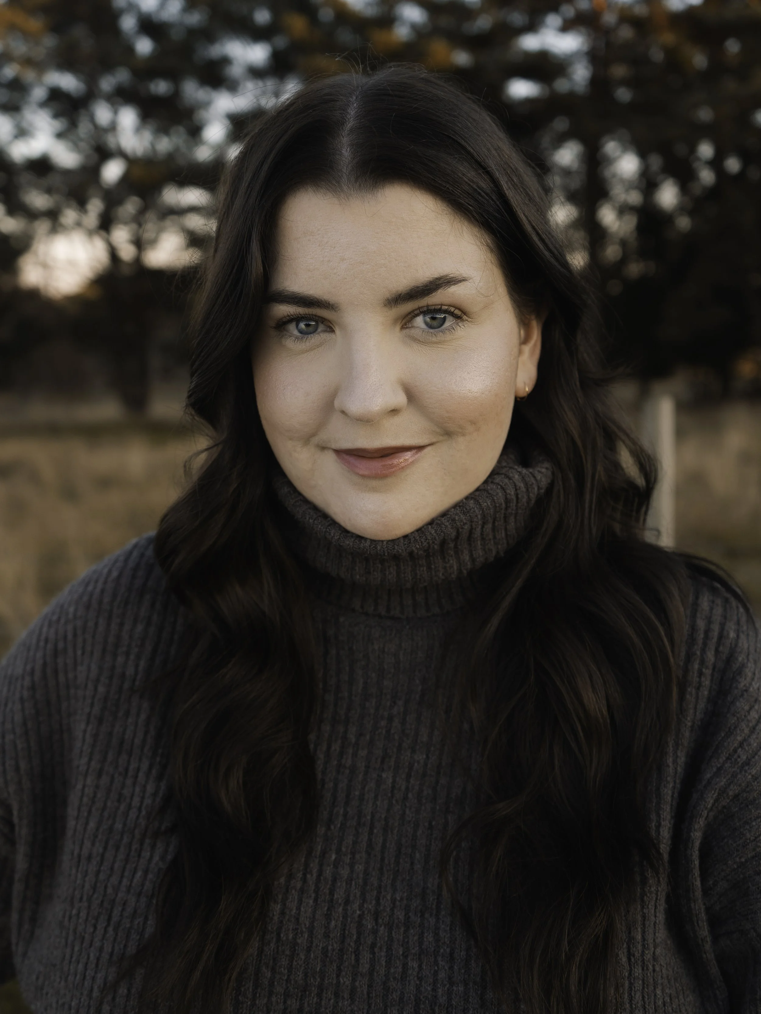 A close-up of a woman with long dark hair, blue eyes, and wearing a dark gray turtleneck sweater, smiling outdoors with trees in the background during autumn.