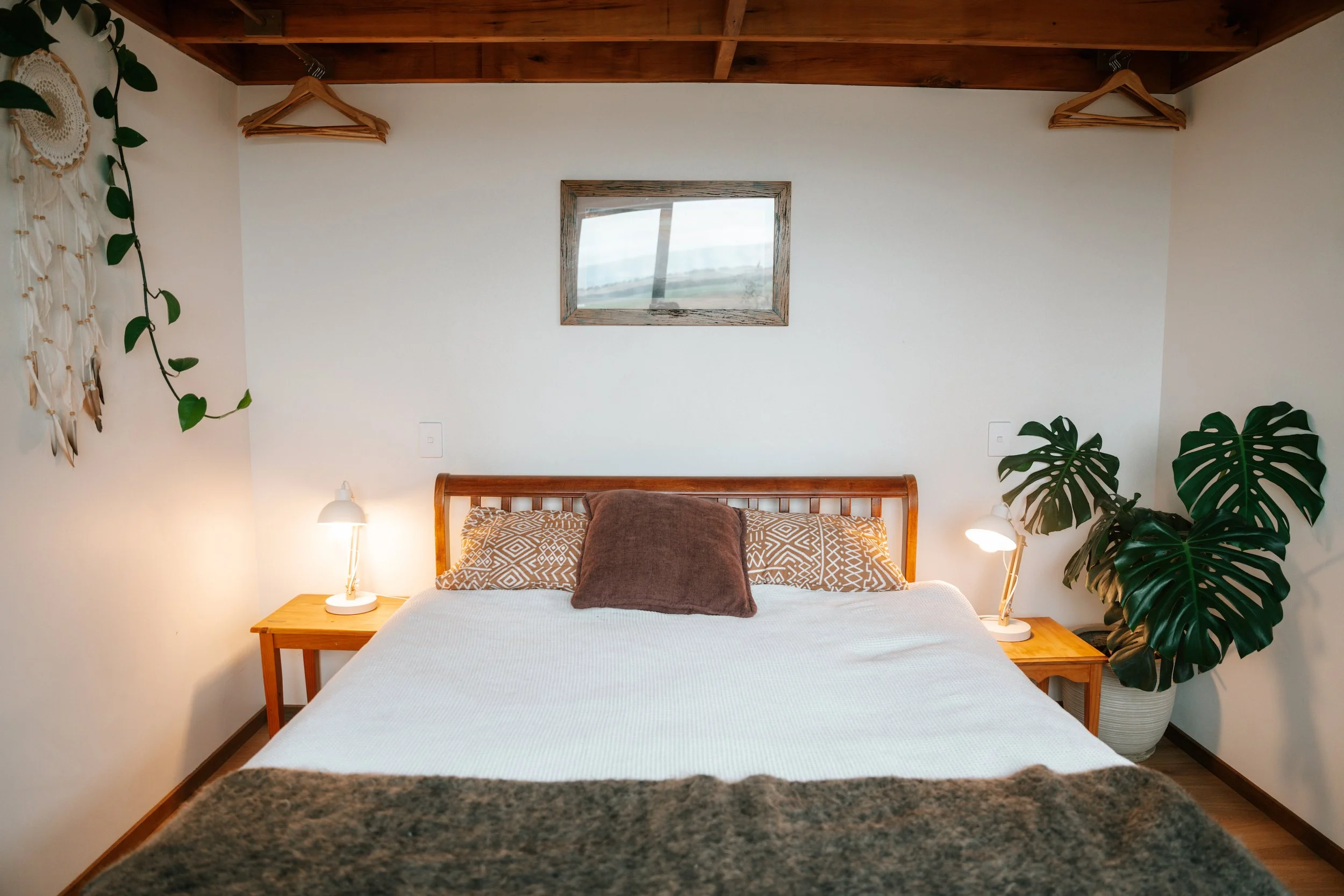 A cozy bedroom with a wooden bed frame, white bedding, and decorative pillows. Two small white lamps sit on wooden bedside tables on each side. A window with a wooden frame is centered on the wall behind the bed, and greenery including a large Monste