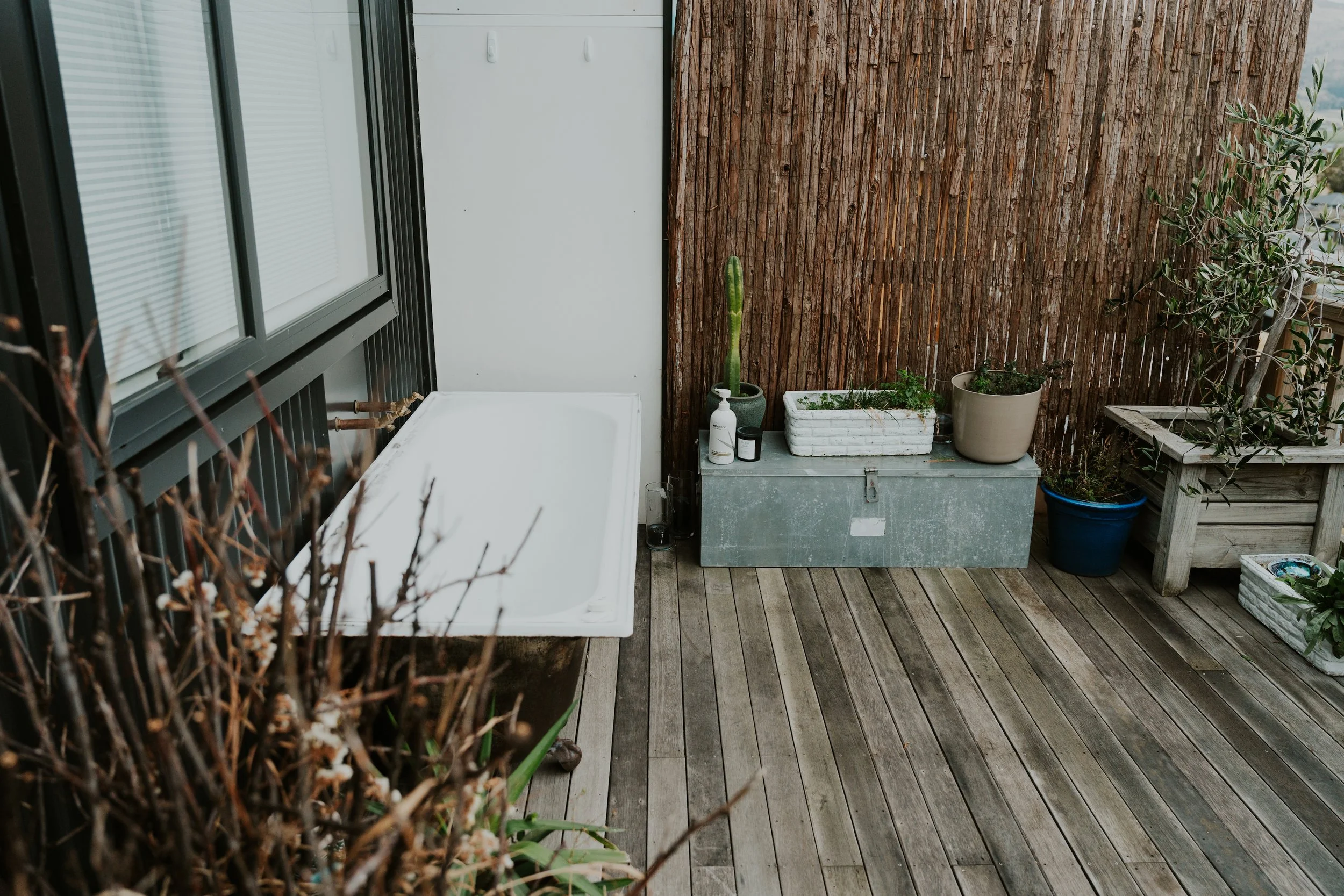 A small balcony with wooden flooring, potted plants, and a wooden privacy screen. There is a white container with a cactus, a white box with plants, and other pots. A window with blinds is on the left.