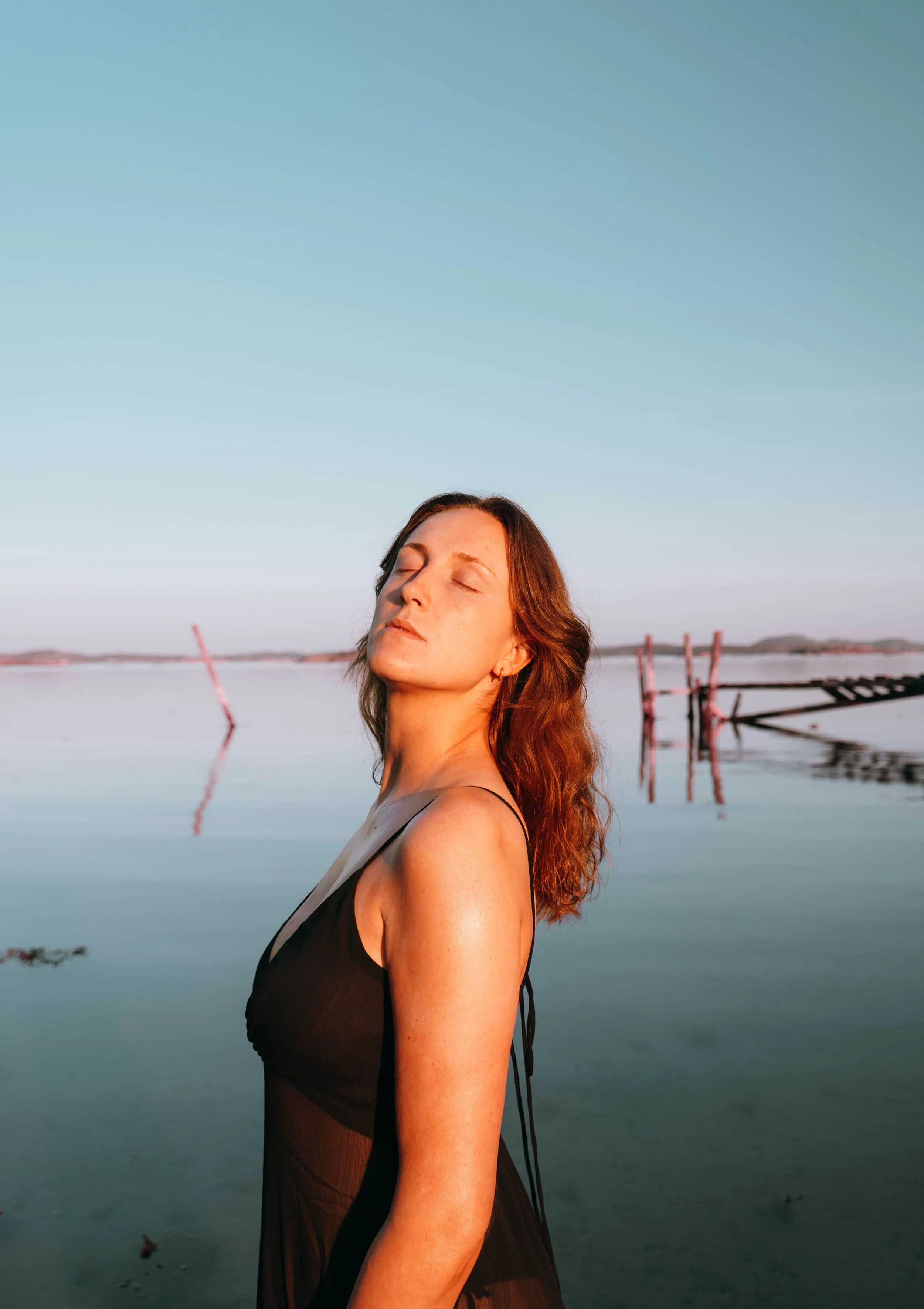 A woman with closed eyes and a serene expression stands near a body of water under a clear sky, with some structures and shoreline visible in the background.