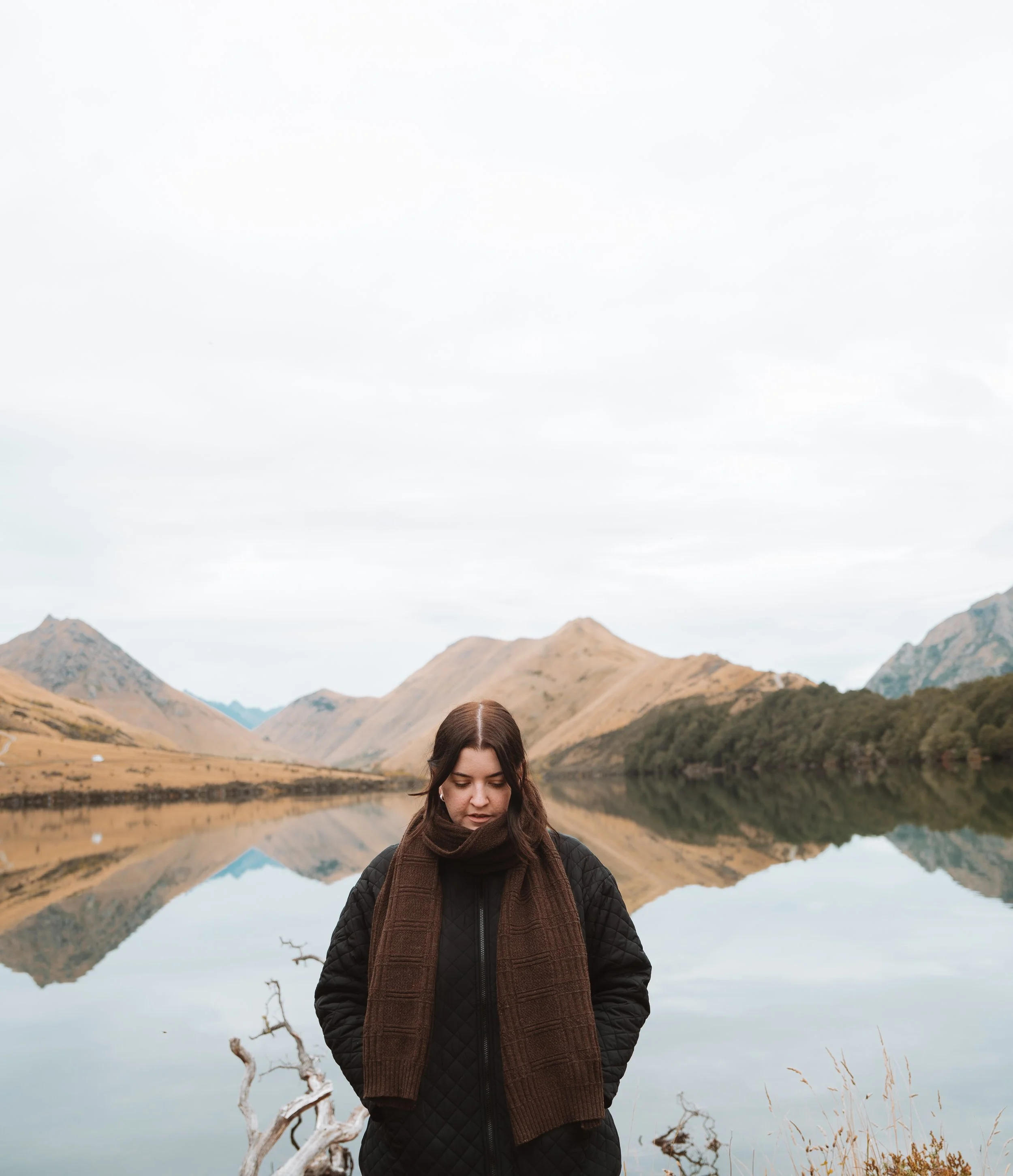A woman with dark hair standing by a lake with mountain scenery, wearing a black quilted jacket and a brown scarf, with reflection of mountains and sky in the water.