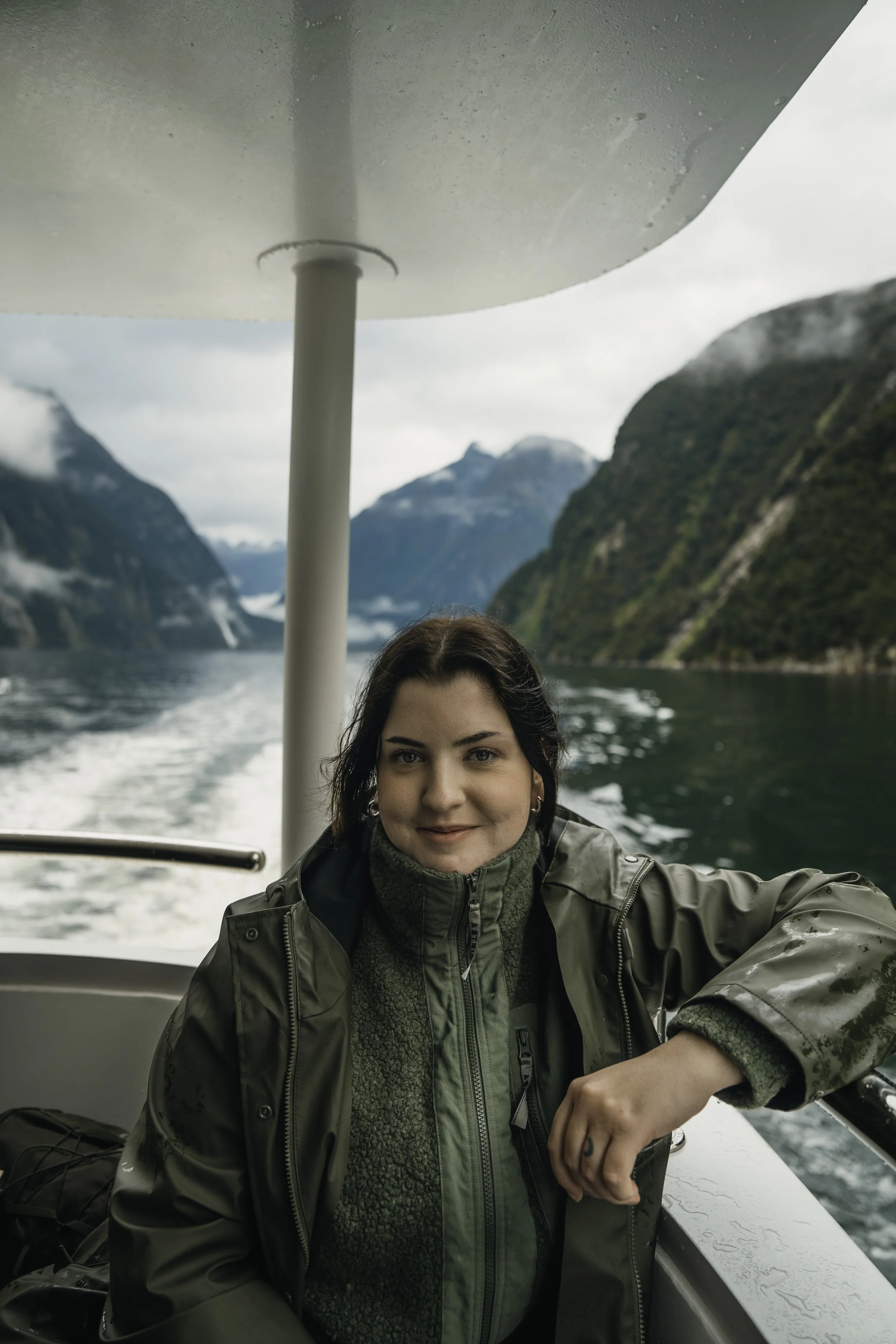 A woman sitting on a boat with a scenic fjord and mountainous landscape in the background, wearing a green rain jacket.