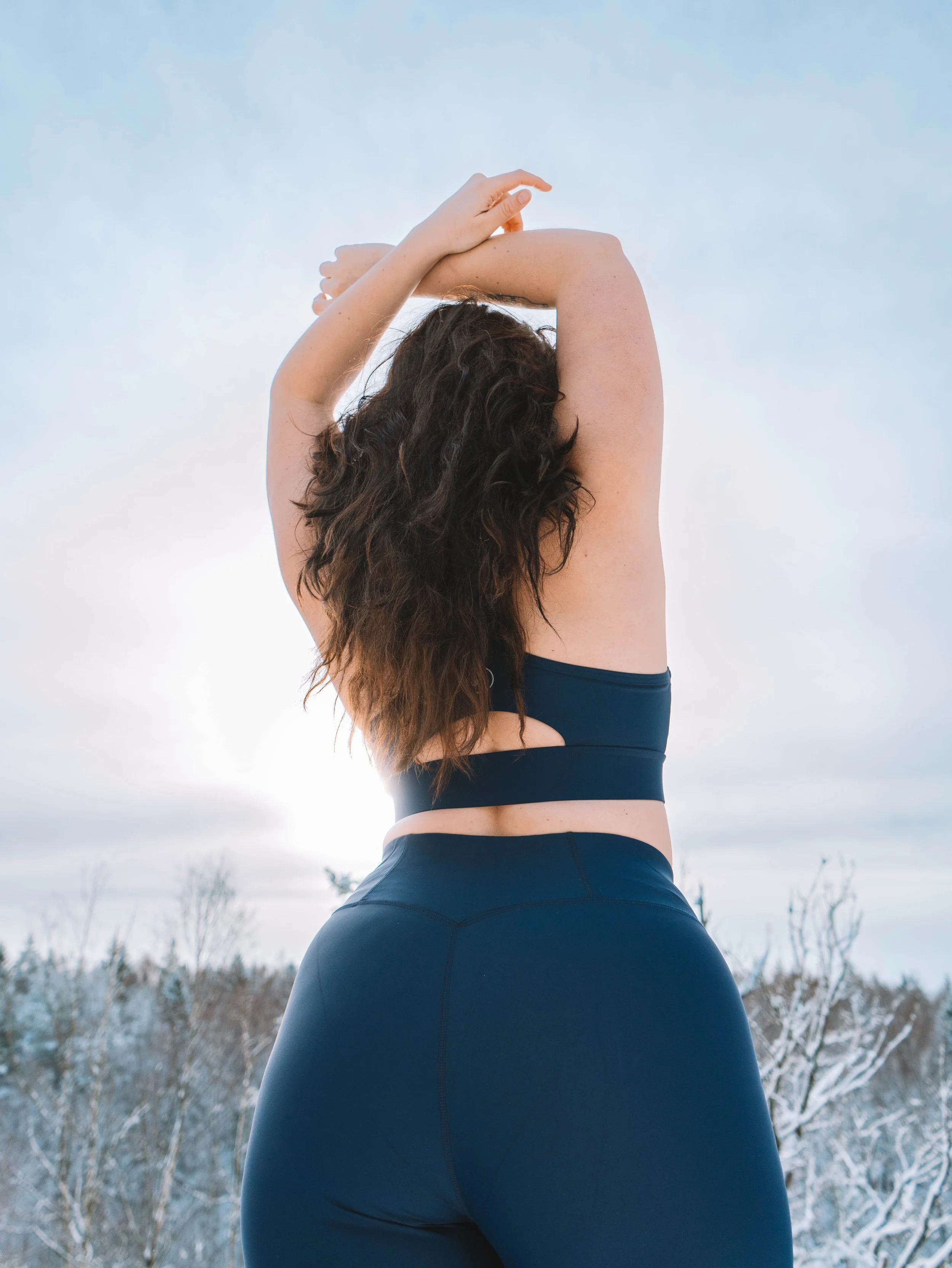 A woman in athletic clothing stretching outdoors with a snowy landscape and trees in the background.