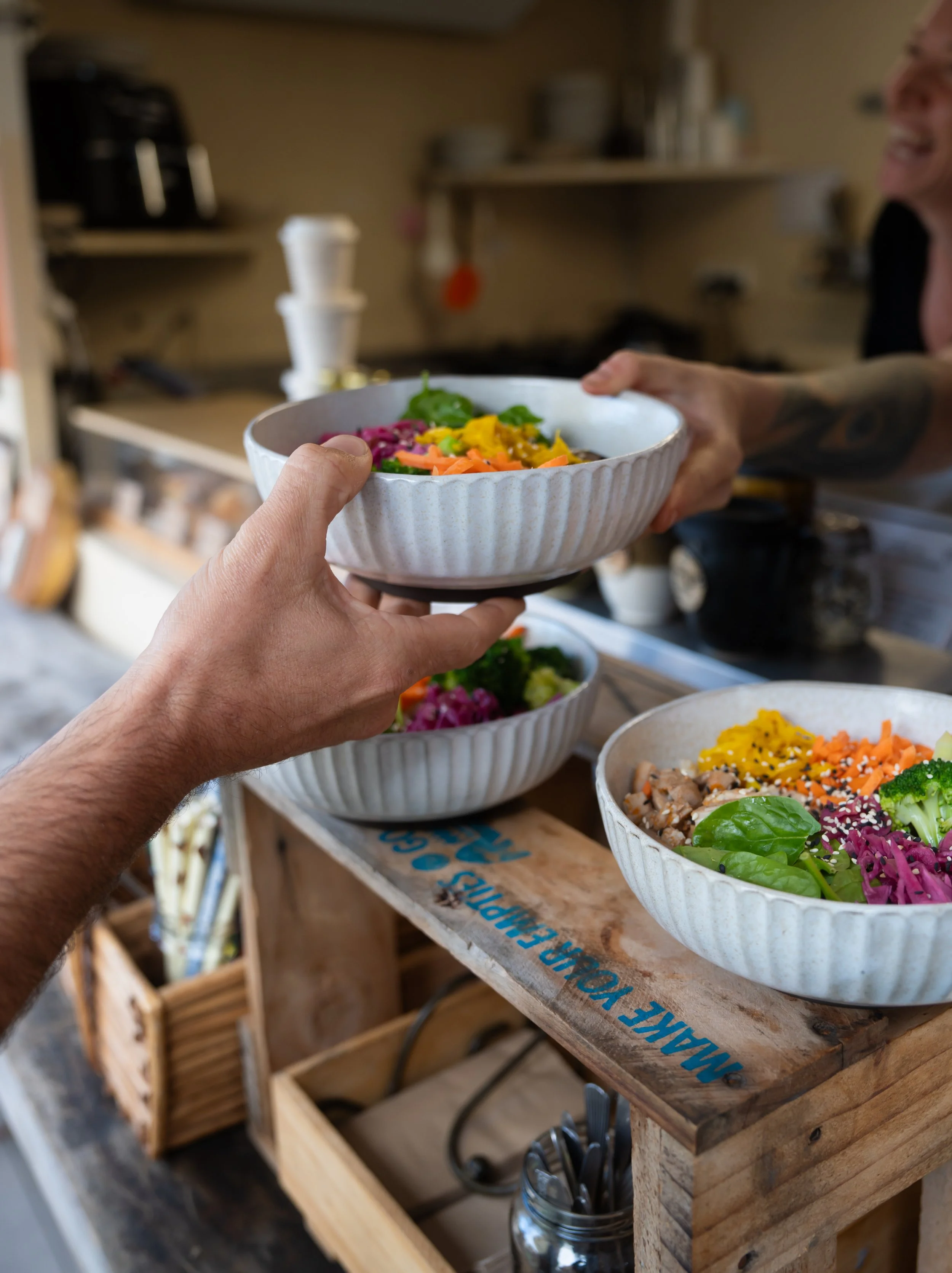 A person handing a bowl of colorful salad to another person in a kitchen, with more bowls of salad on a rustic wooden shelf.