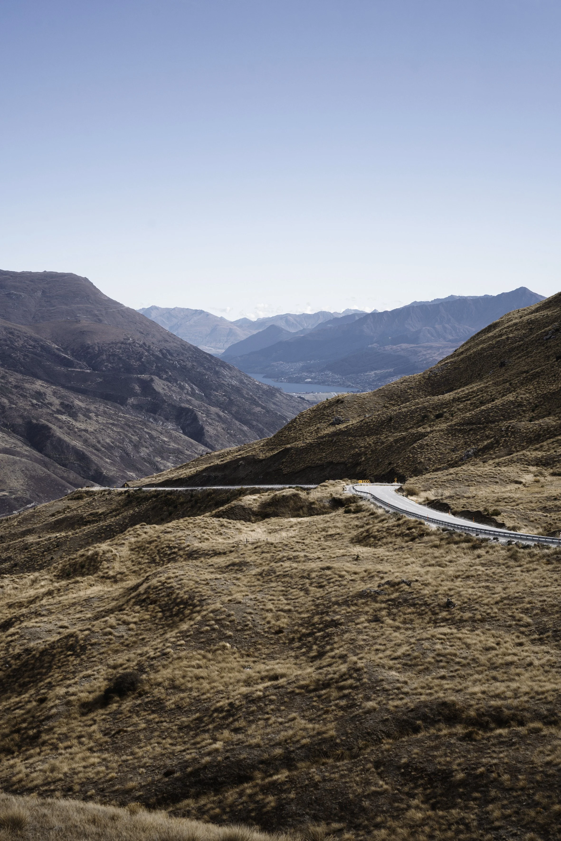 A mountain landscape with dry brown and green hills, a winding road, distant mountains, and a blue sky