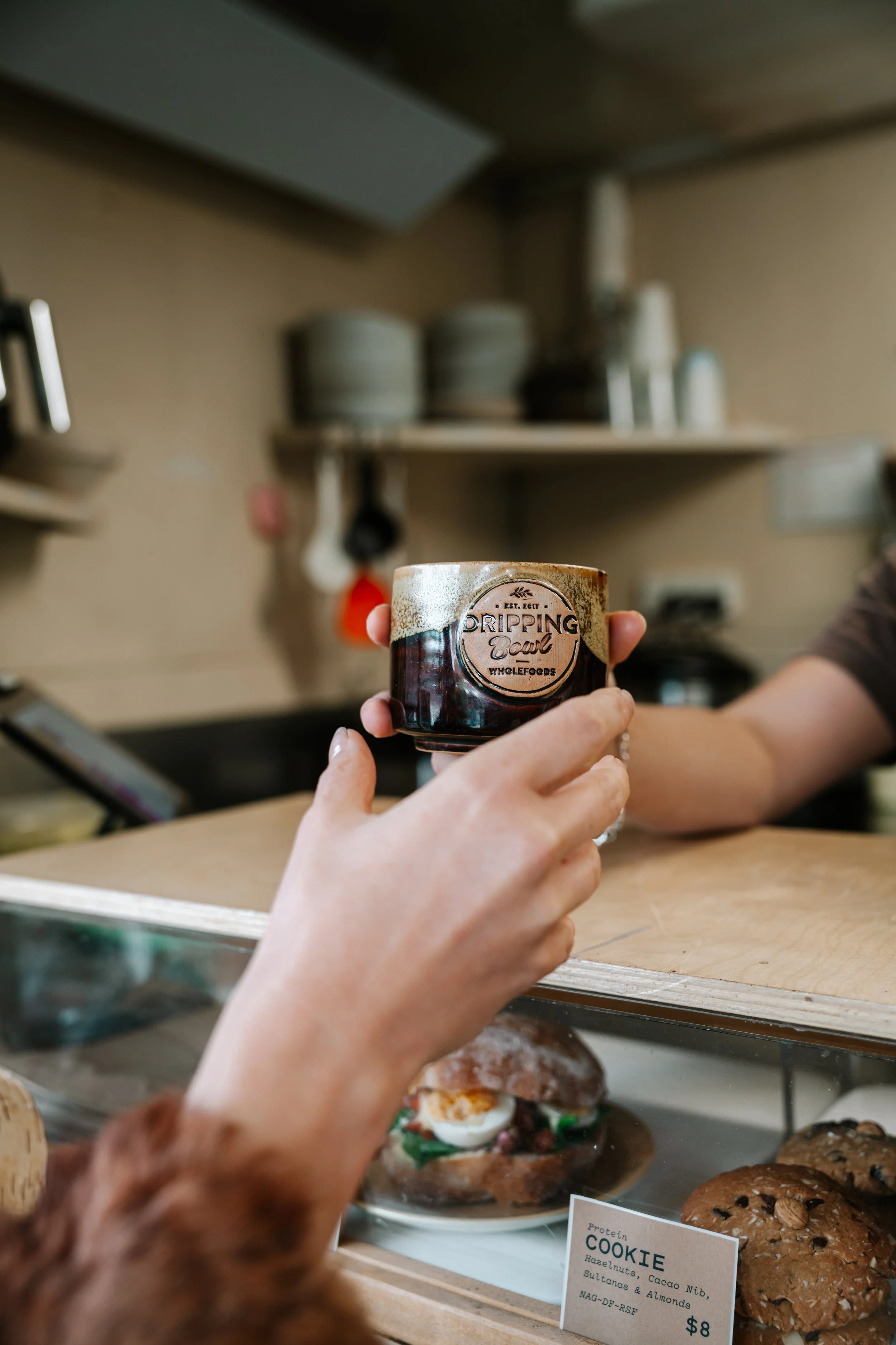 Person holding a small jar labeled 'Dripping Sauce' at a bakery display with assorted baked goods, including cookies, behind the glass case.
