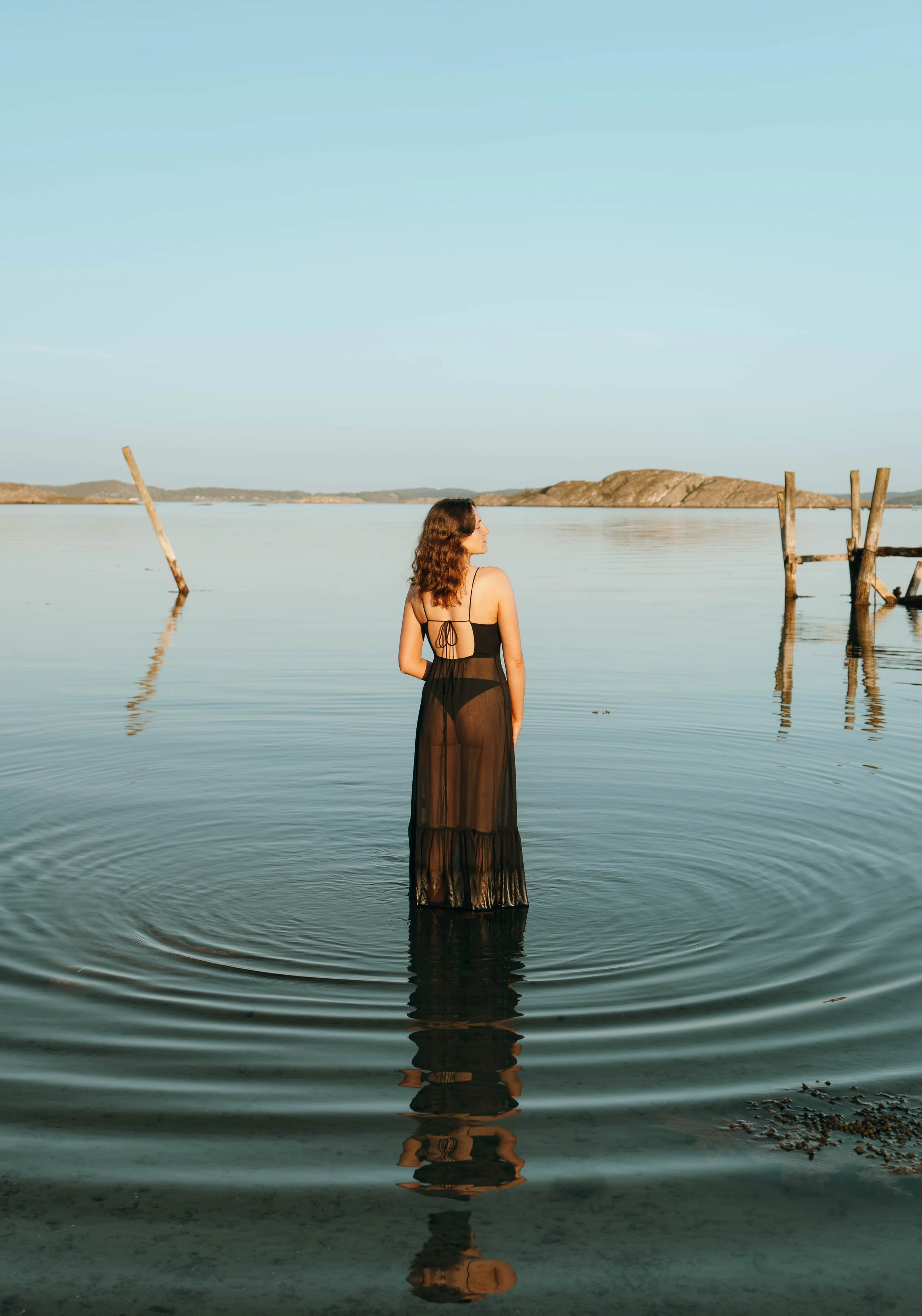 Woman with wavy brown hair in a black sheer dress standing in calm water near the shoreline, with land and hills in the background and a partly cloudy sky.
