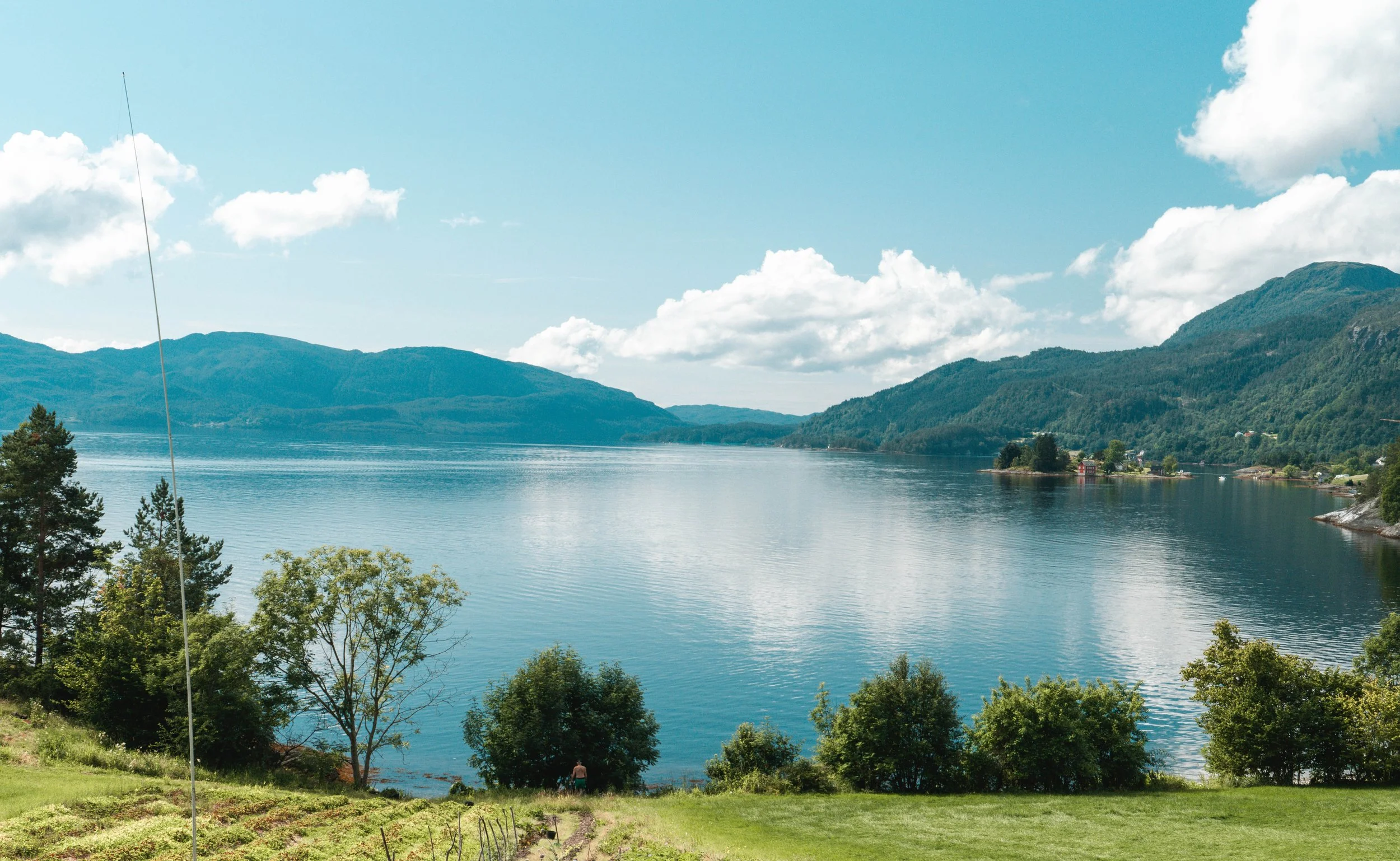 A scenic view of a river surrounded by mountains with greenery and trees along the riverbank, under a partly cloudy blue sky.