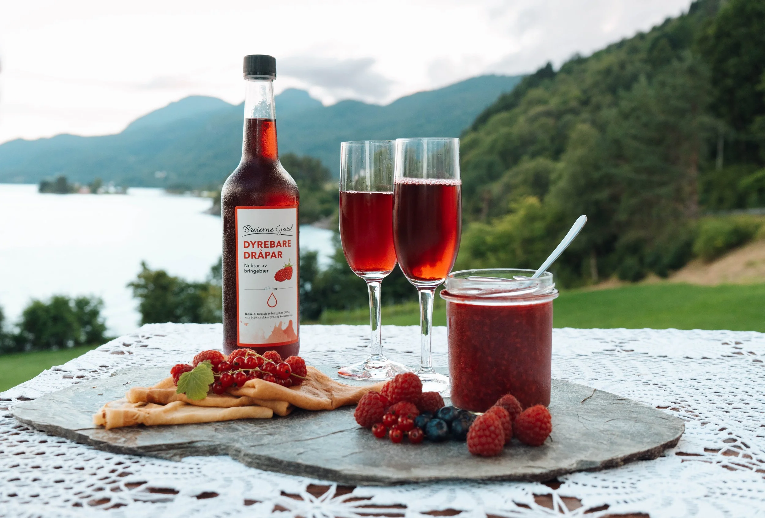 Outdoor table with a bottle of berry syrup, two glasses filled with berry syrup, a jar of berry jam, fresh berries, and a plate of waffles with berries, set against a scenic mountain and lake background.