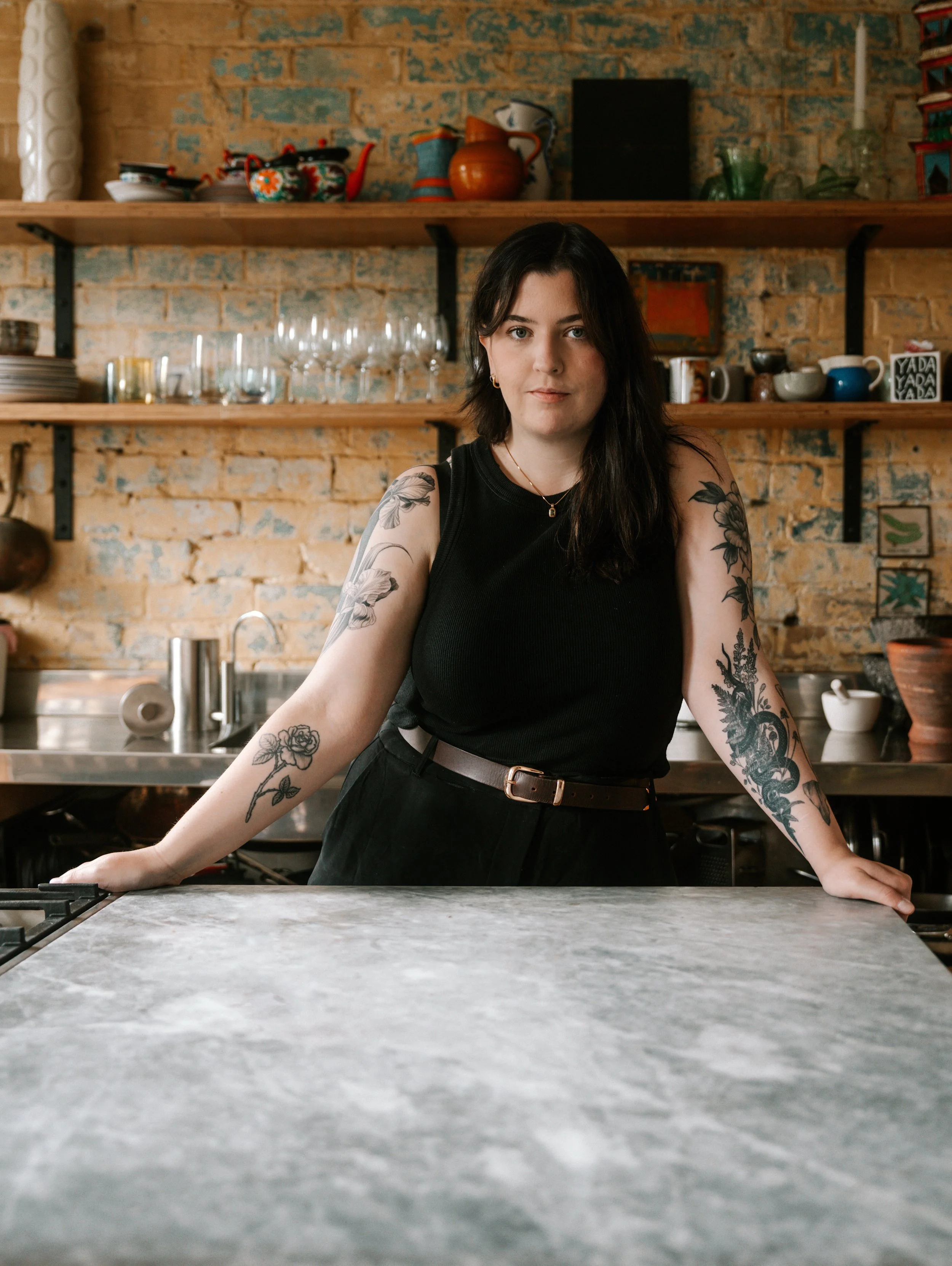 A young woman with dark hair and tattoos on her arms sits at a kitchen counter, facing the camera. Behind her are open wooden shelves with dishes, glasses, and decorative items on a brick wall.