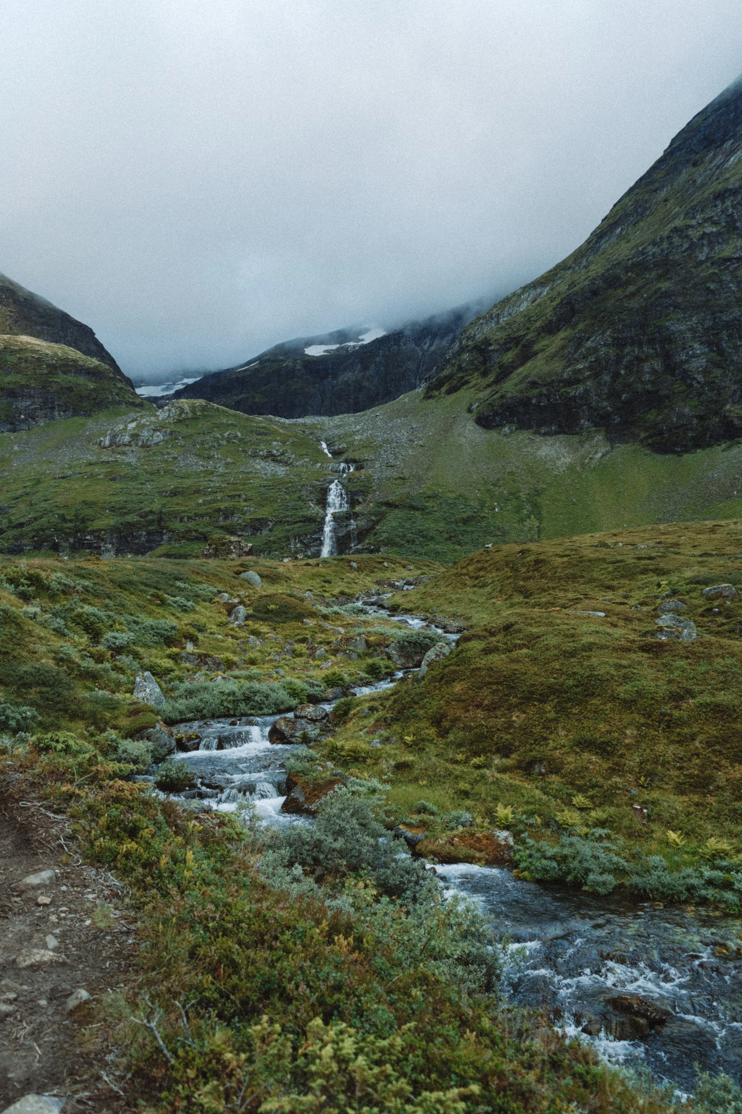 Mountain valley with a small waterfall flowing through green vegetation and rocky terrain, surrounded by steep, moss-covered mountains and foggy sky.