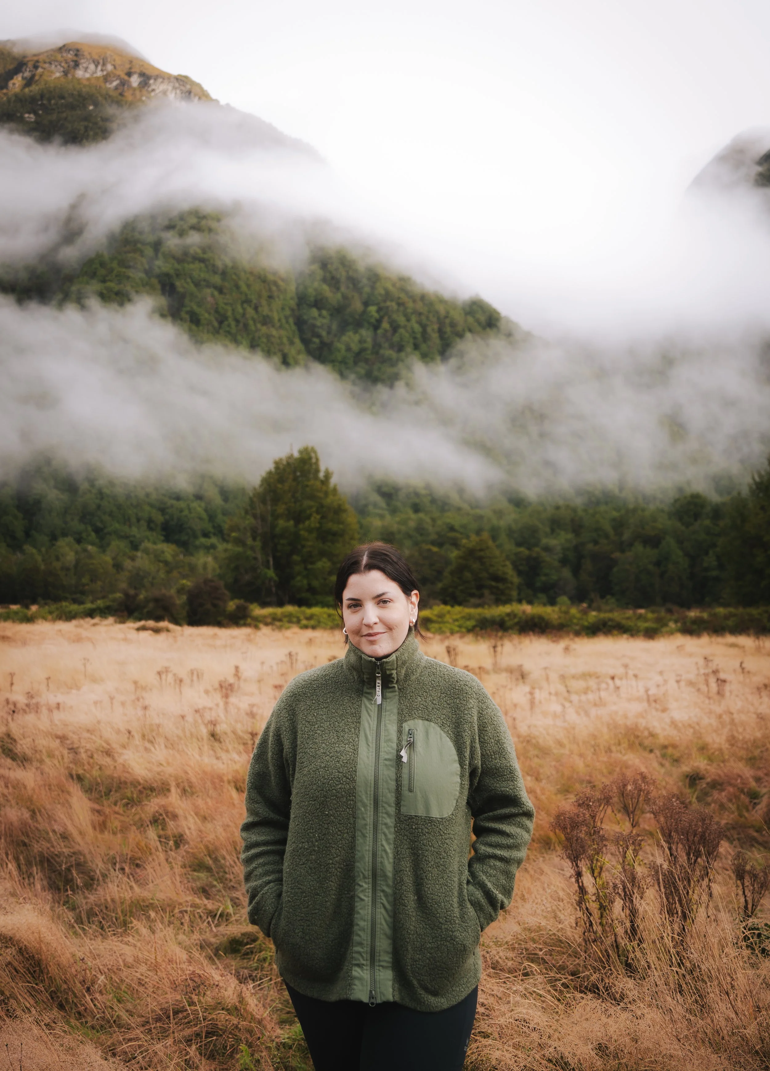 A young woman with dark hair and light skin is standing in a field of tall, dry grass. She is wearing a green fleece jacket with a zipper pocket and black pants. Behind her are green trees and a mountain shrouded in mist or clouds.