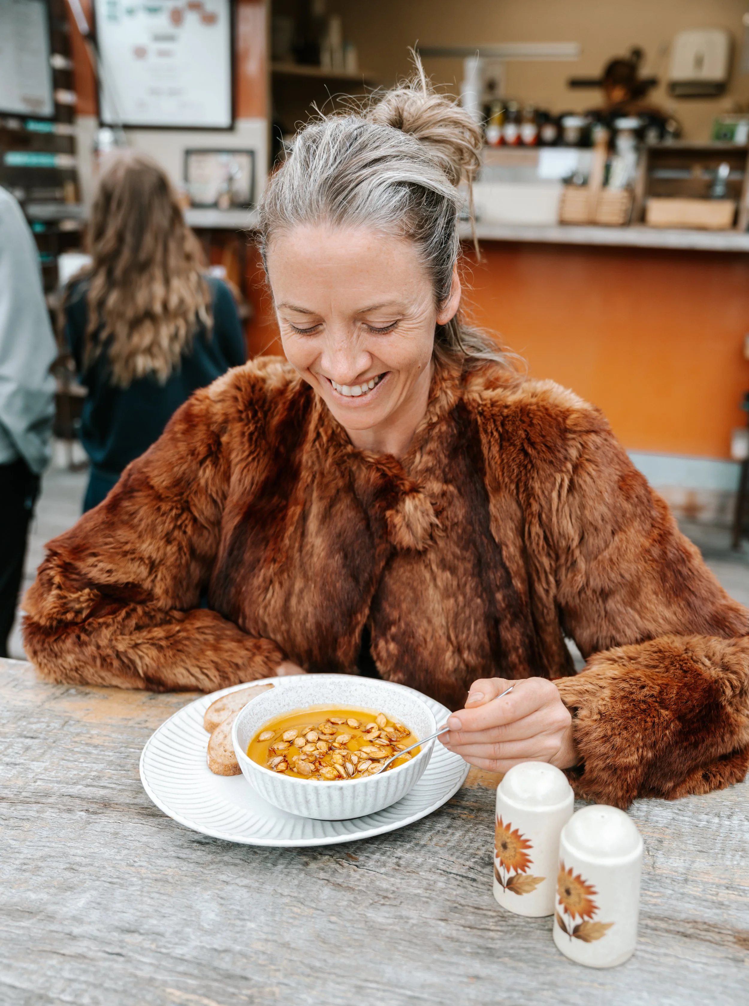 A woman with gray and blonde hair in a messy bun, wearing a faux fur coat, is smiling while sitting at a rustic wooden table in a cafe. She is holding a spoon and about to eat a bowl of pumpkin seed soup garnished with pumpkin seeds. There are salt and pepper shakers on the table, along with some bread slices. The background shows other patrons and cafe decor.