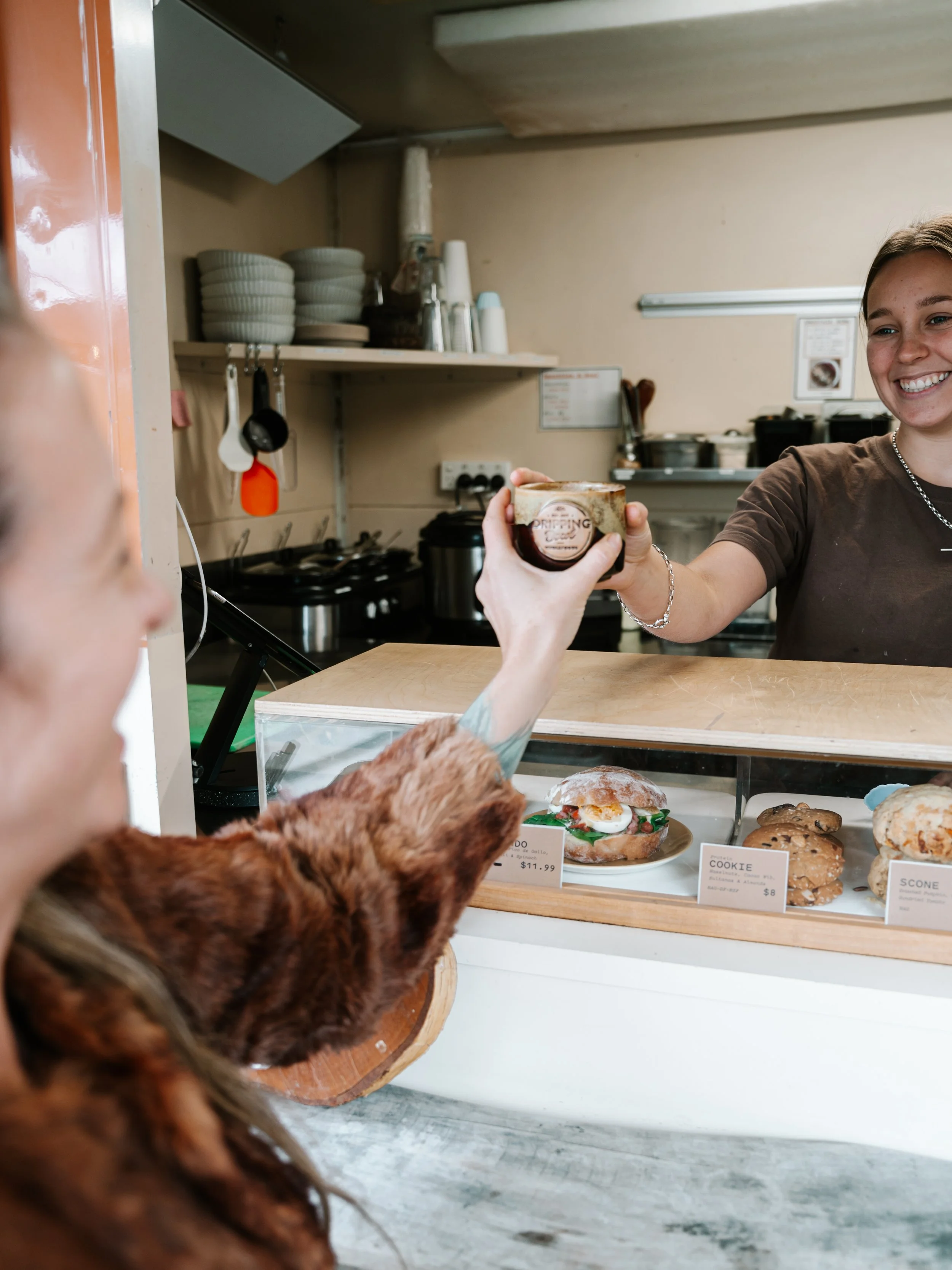 A person handing a small cup of coffee or dessert to a smiling employee at a bakery or cafe counter. The counter displays assorted baked goods, including a sandwich and cookies. Shelves in the background hold stacked bowls and cups, with kitchen uten