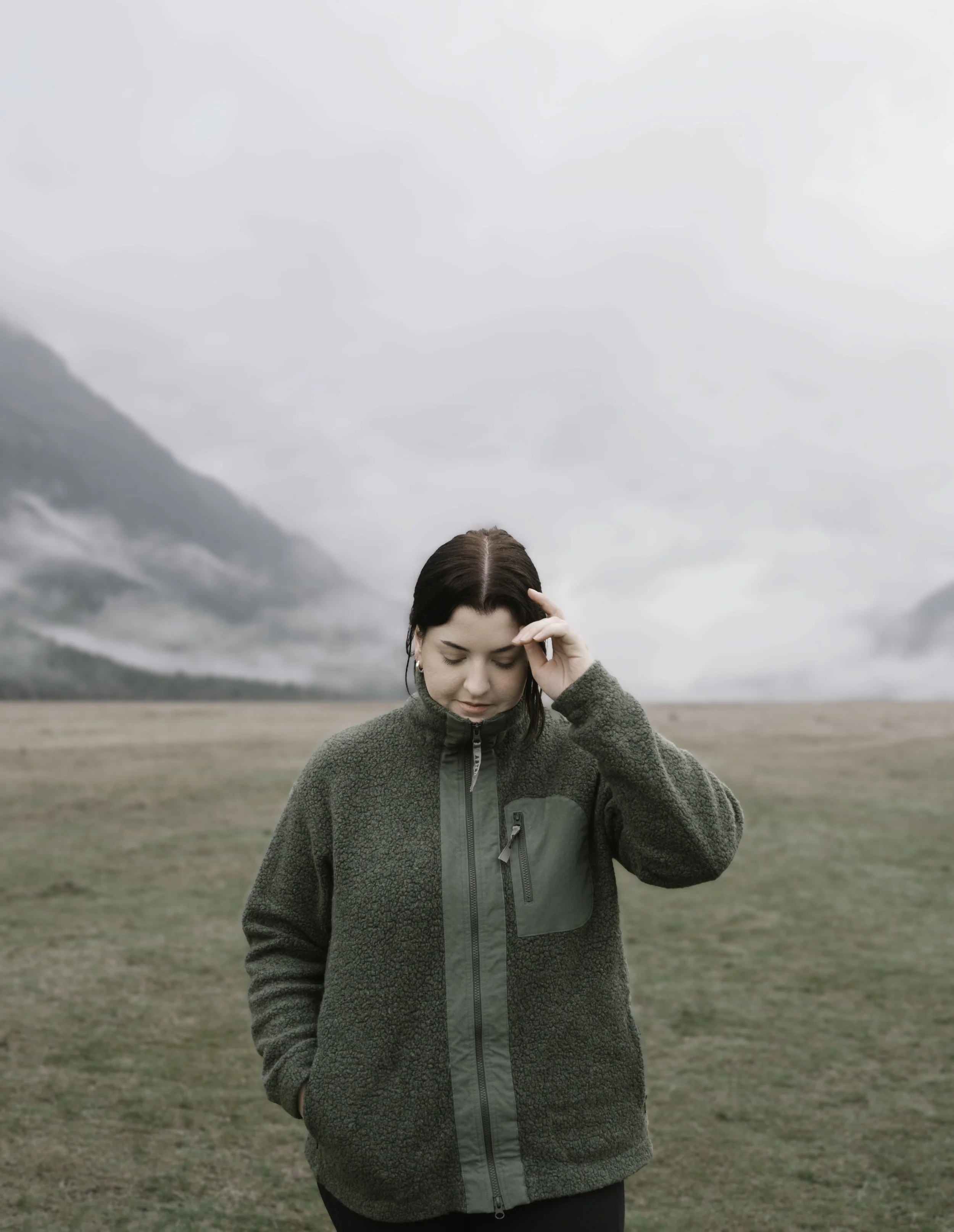 A young woman with brown hair in a green fleece jacket standing in an open field with mountains and cloudy sky in the background.
