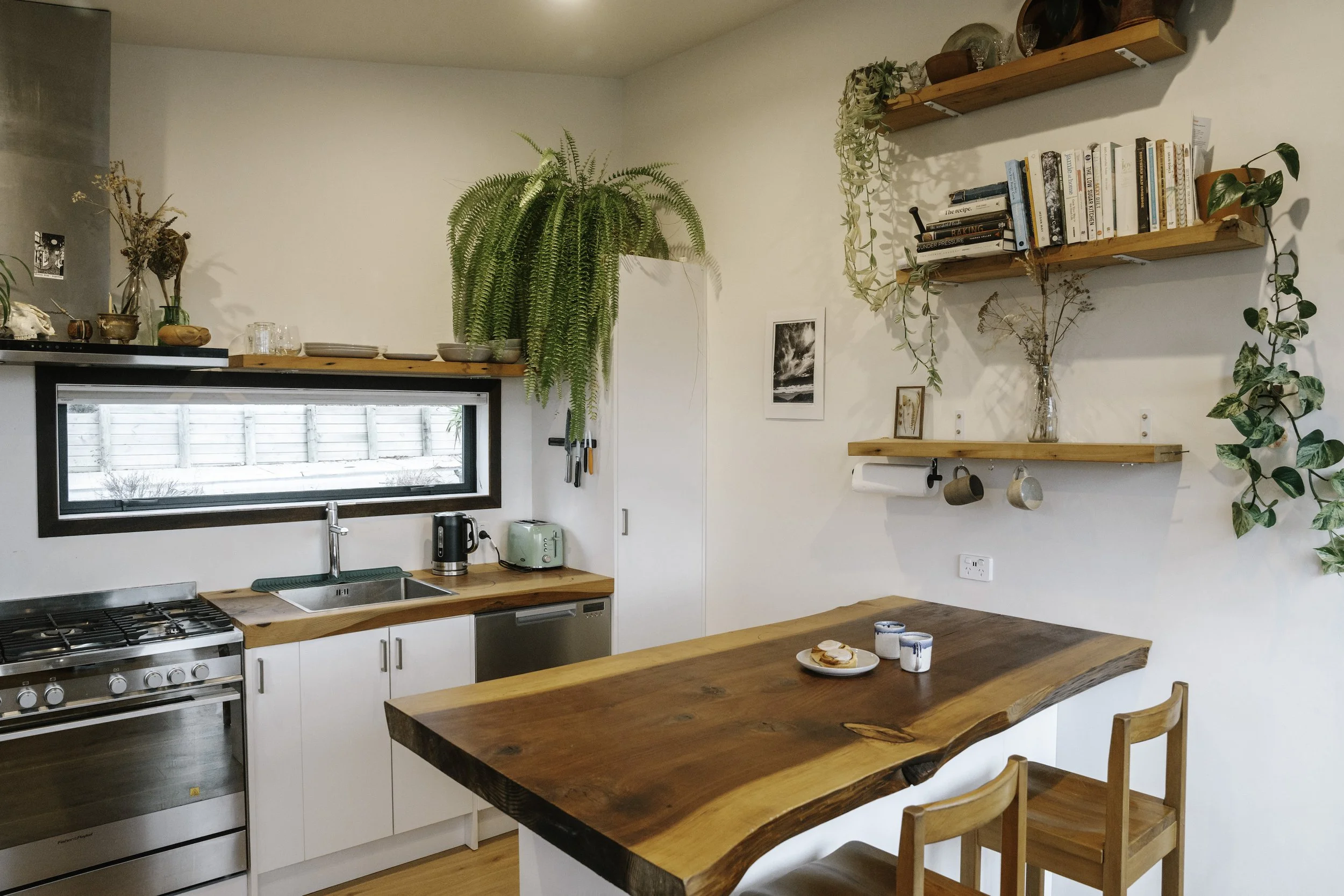 A cozy kitchen with white cabinets, a wooden countertop, and a kitchen window. Decor includes potted plants, shelves with books, and small vases. There is a gas stove, kettle, toaster, dish rack, and a paper towel holder. A wooden dining table with c