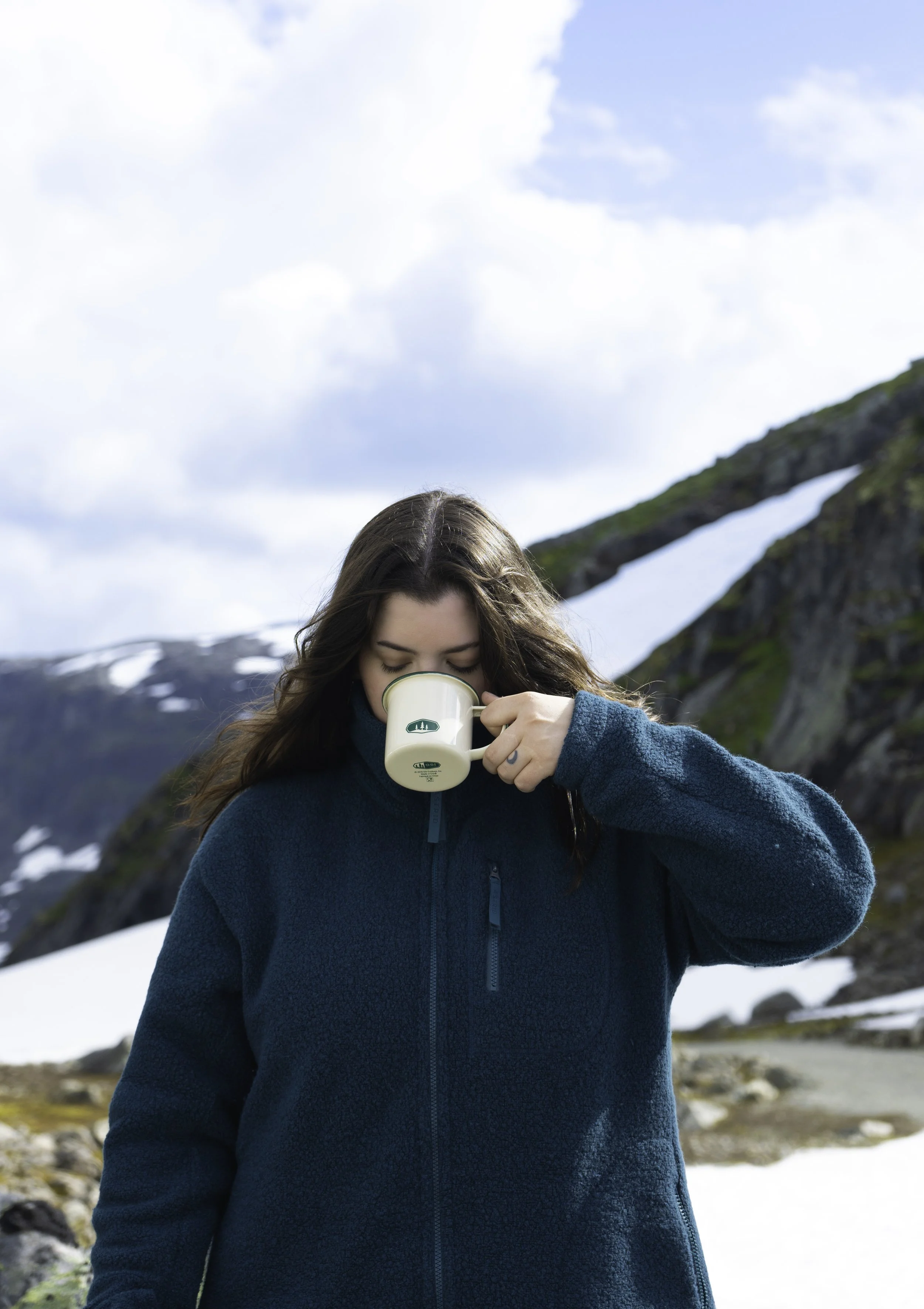 A woman with long brown hair wearing a teal fleece jacket, drinking from a beige mug outdoors in a mountainous area with snow and cloudy sky.