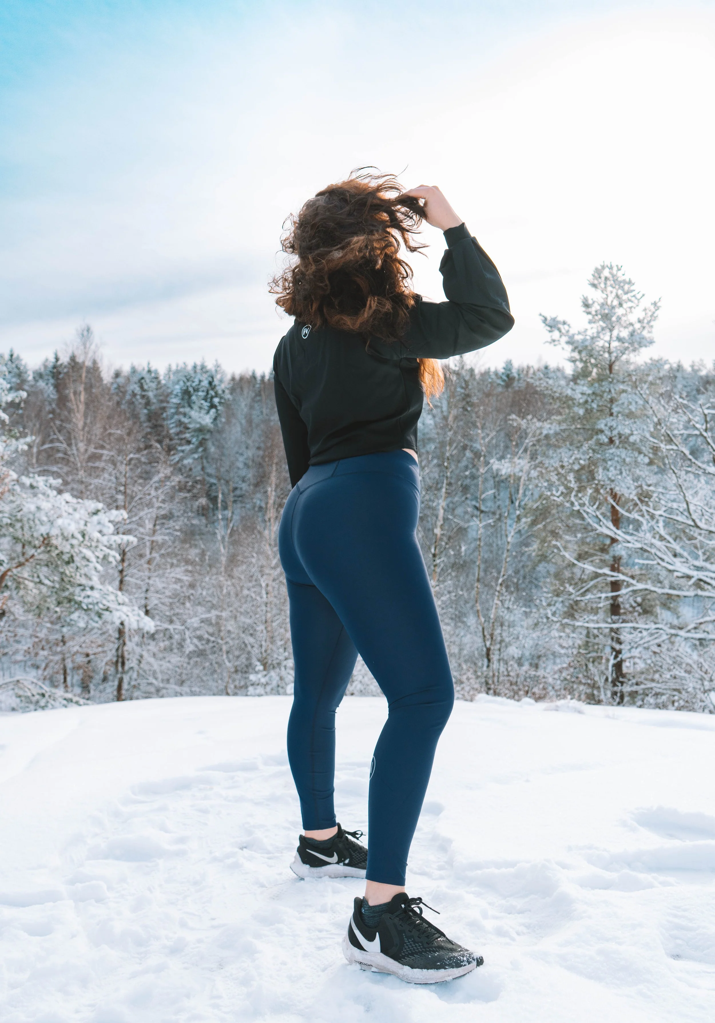 A woman with curly hair standing outdoors on snow, wearing black athletic clothes and black Nike running shoes, with a snowy forest in the background.